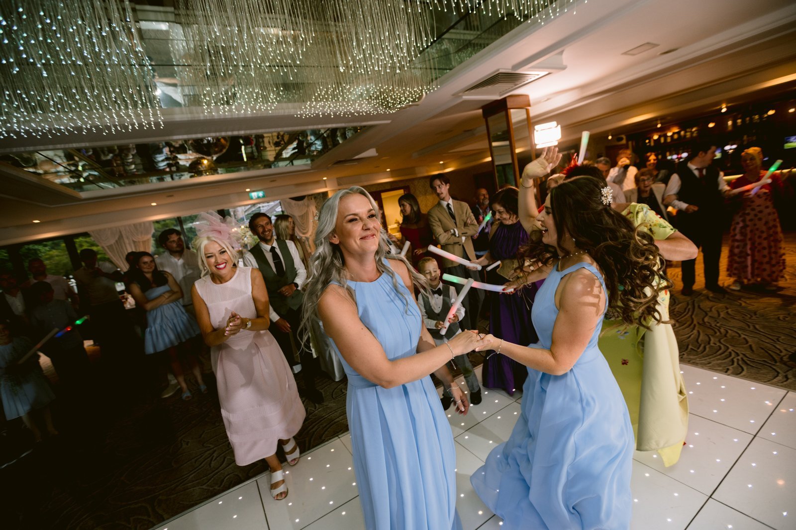 A group of people in formal attire dance and celebrate on a lighted floor under string lights at an August Summer Wedding at Armathwaite Hall.
