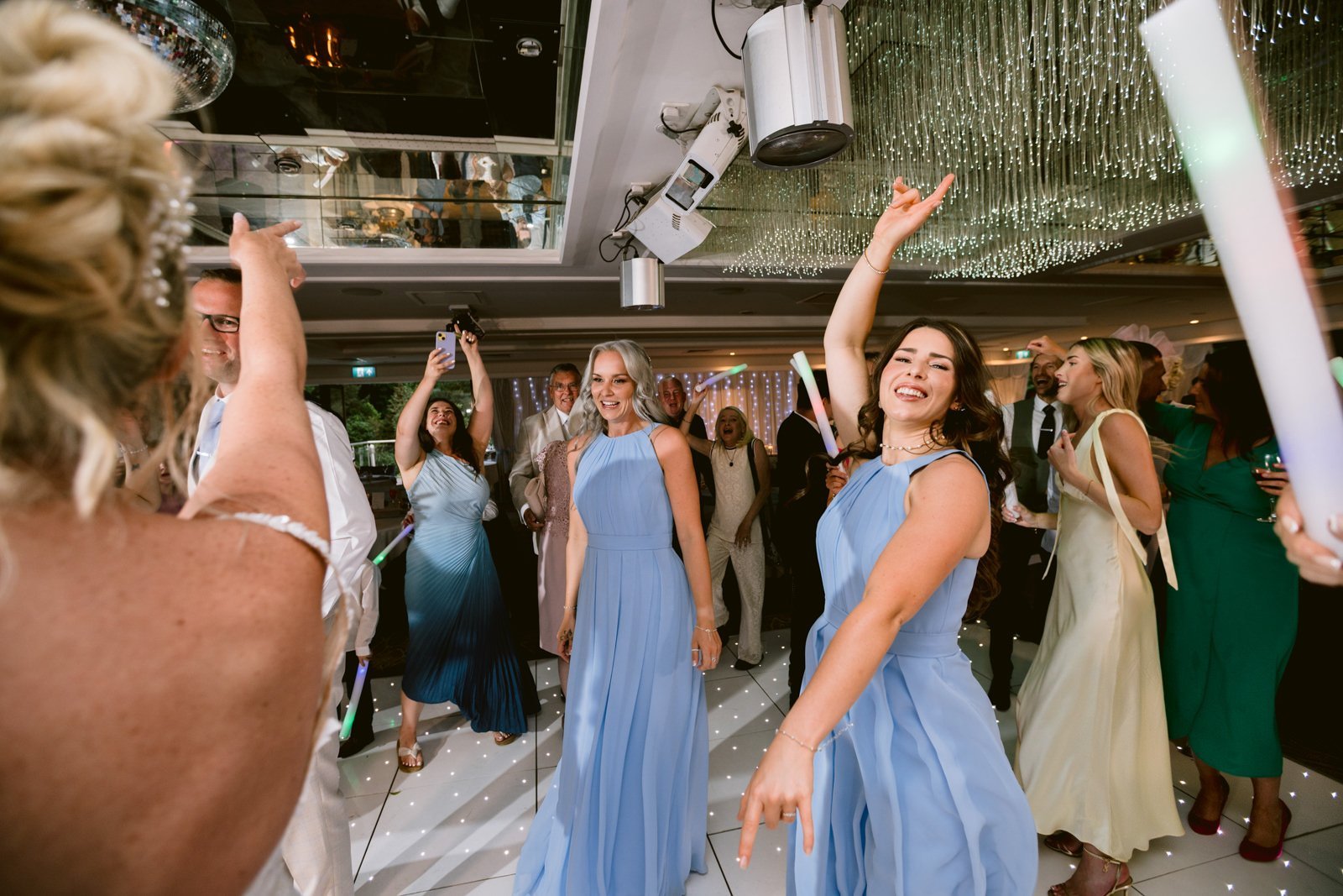A group of people, some in light blue dresses, dance and celebrate on a white-lit floor at an indoor August Summer Wedding at Armathwaite Hall, with glowing hanging lights above.