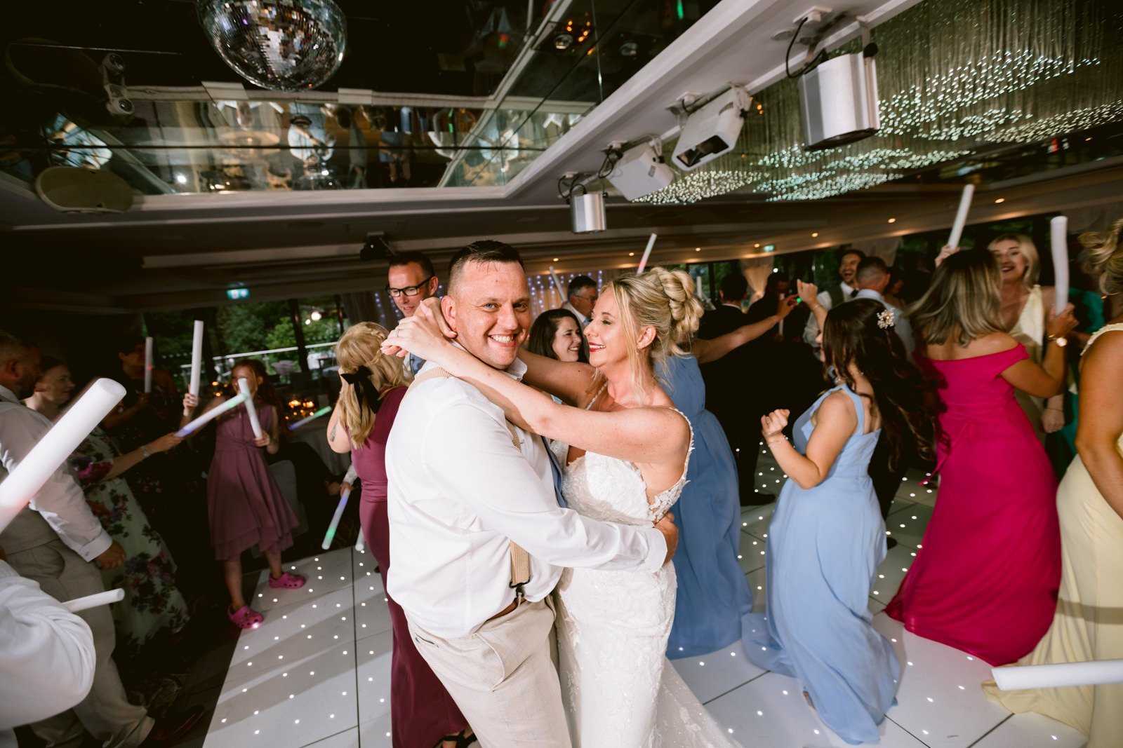 A bride and groom dance closely on a lit-up floor at their lively August wedding, surrounded by guests in colorful dresses and suits, celebrating at beautiful Armathwaite Hall.