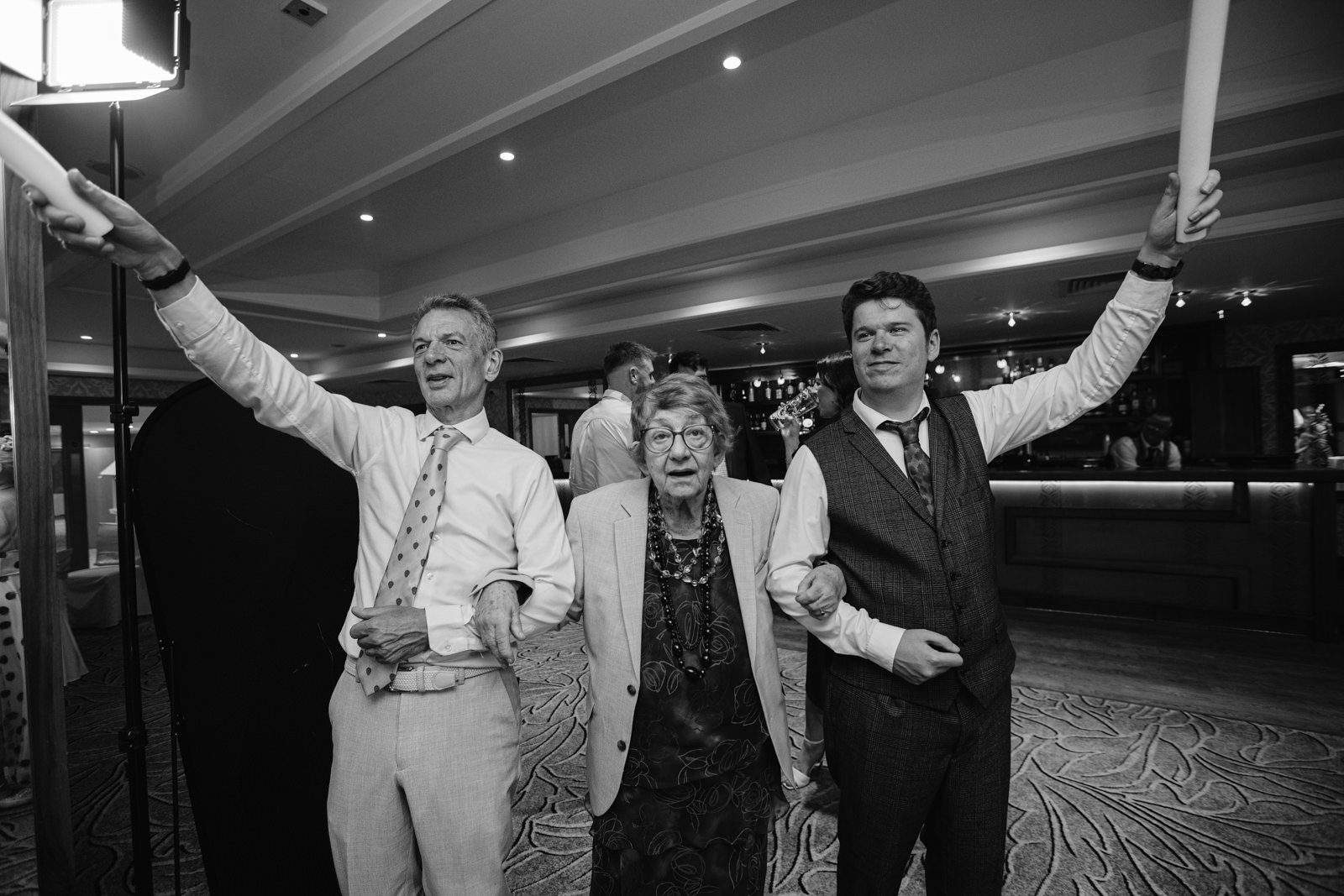 Three people dressed formally stand together indoors at Armathwaite Hall; two men hold up long objects while an elderly woman links arms between them. The setting suggests a joyful August Summer Wedding social event.