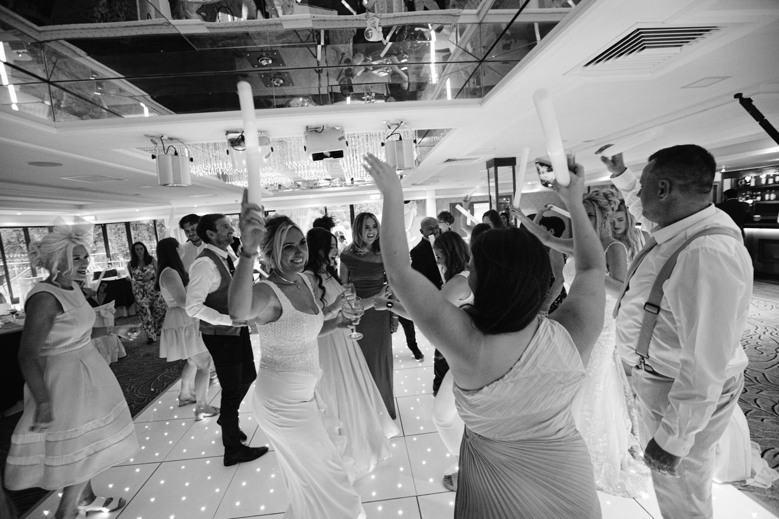 A group of people in formal attire dance and hold glow sticks on a lit dance floor at an indoor August summer wedding reception at Armathwaite Hall.