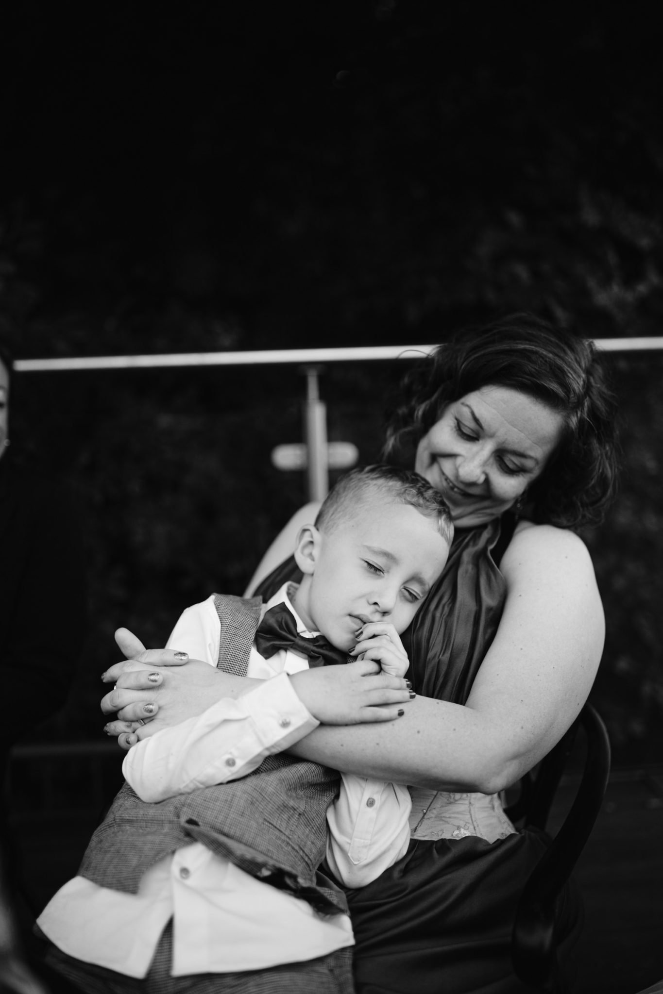 A woman sits and embraces a young boy wearing a vest and bow tie during an August Summer Wedding at Armathwaite Hall. The boy appears calm, resting his head on her arm.