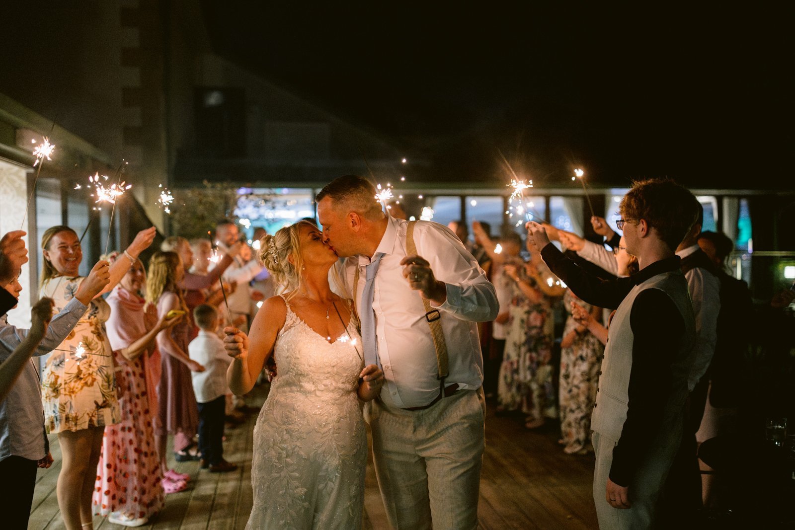 A bride and groom kiss while walking through a crowd of guests holding sparklers at night during their August summer wedding at Armathwaite Hall.