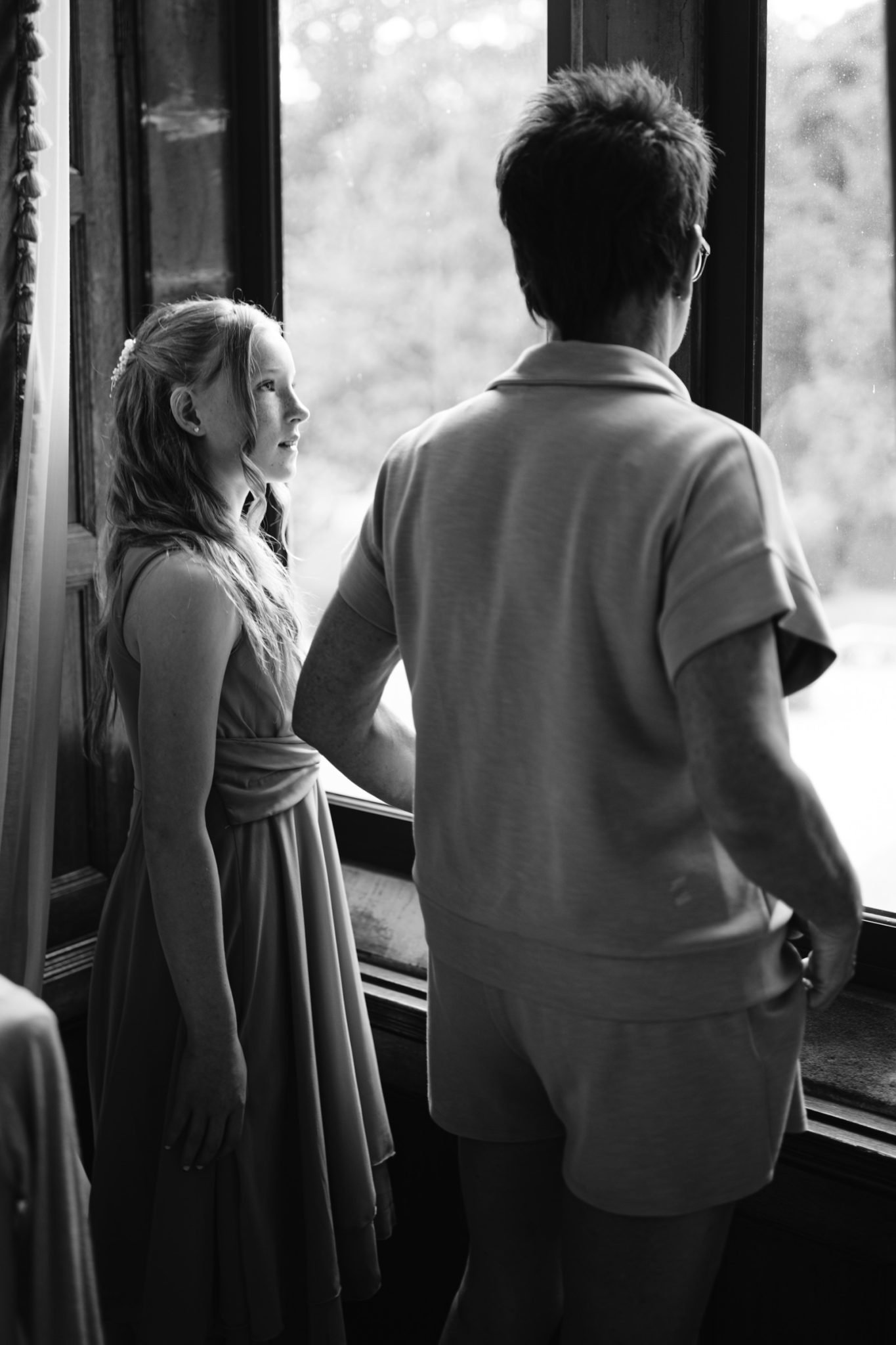 A girl in a dress and an older person in casual clothes stand by a window at Armathwaite Hall, looking outside. The black and white image captures a quiet moment from an August summer wedding.