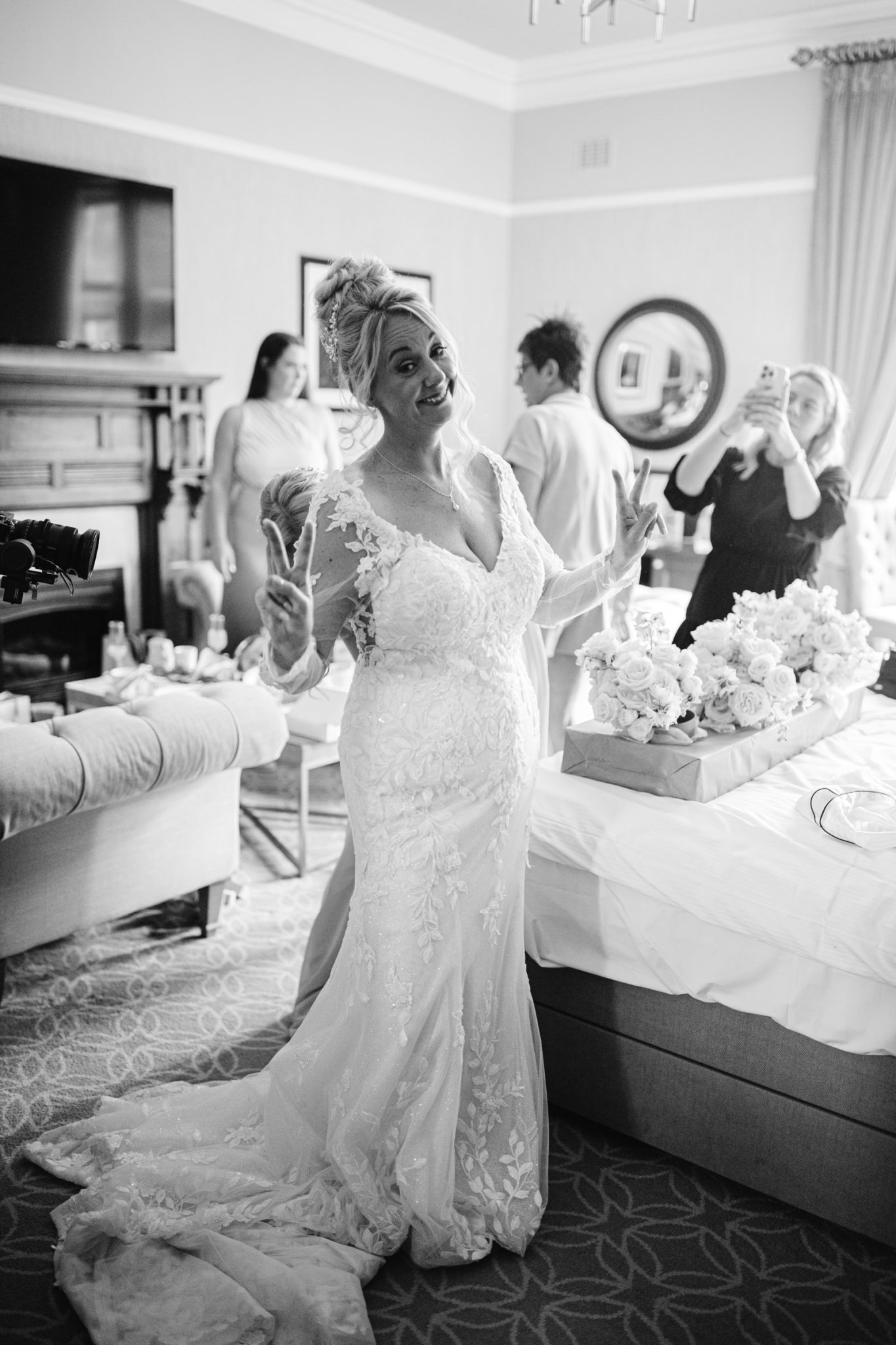 A bride in a lace wedding dress makes peace signs with both hands at her August Summer Wedding, standing in a room at Armathwaite Hall with several people and a bouquet of flowers on the bed.
