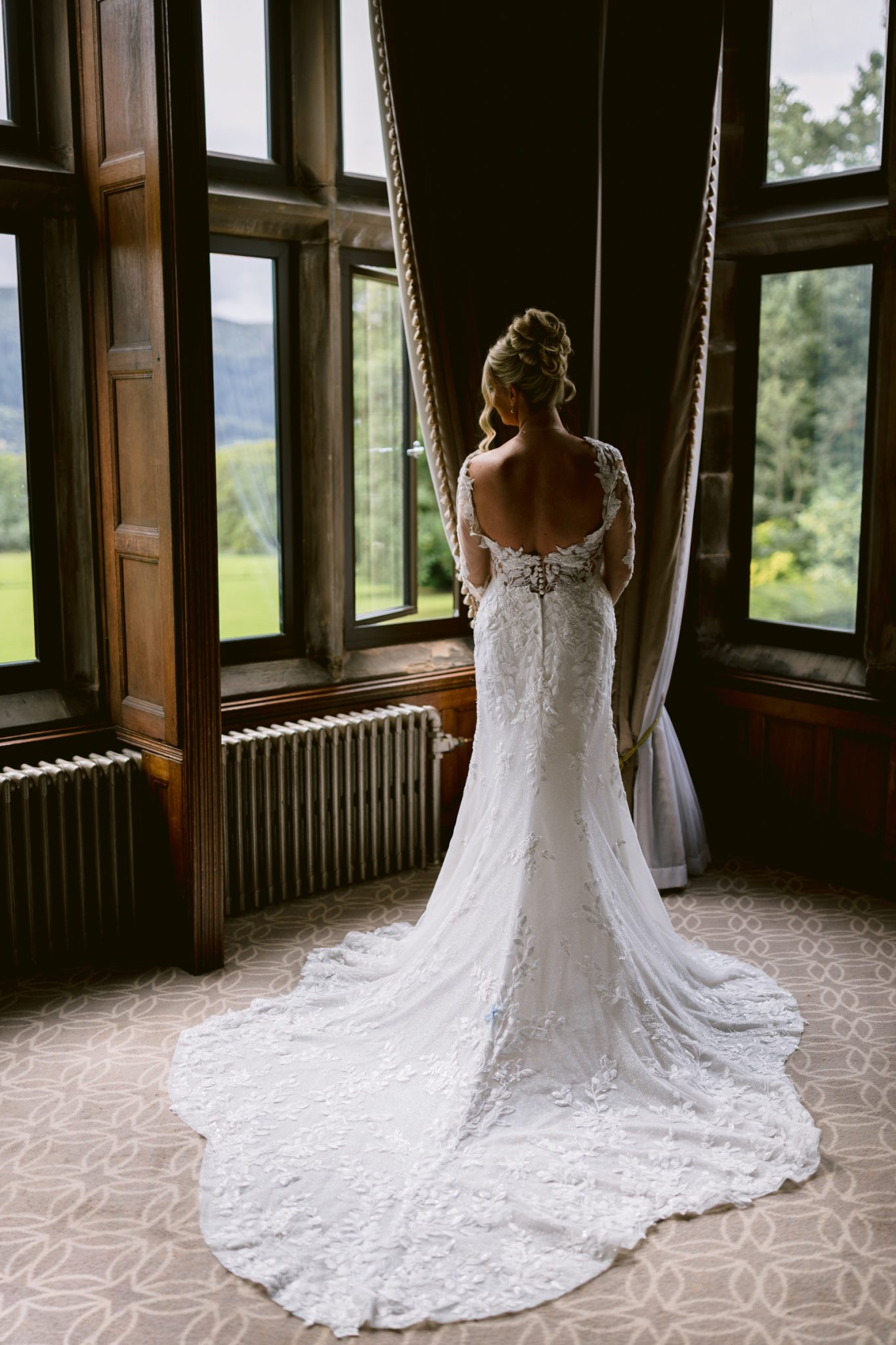 A bride in a white lace wedding dress stands facing large windows at Armathwaite Hall, looking outside on her summer wedding day.