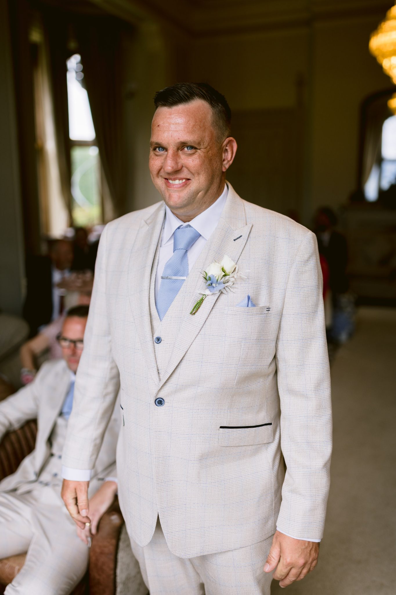 A man in a light-colored checked suit with a blue tie and boutonniere stands indoors, smiling at the camera during an August summer wedding at Armathwaite Hall.