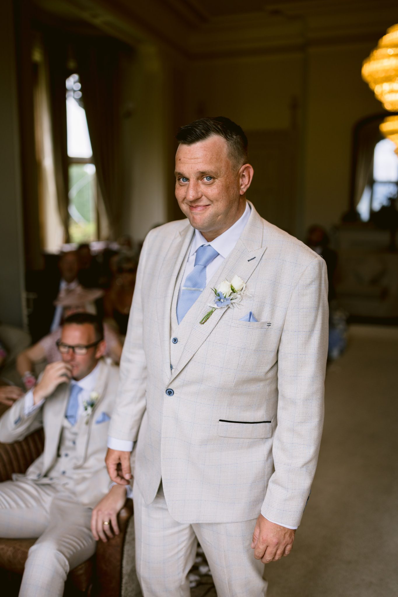 A man in a light-colored suit with a blue tie and boutonniere stands indoors at an August Summer Wedding at Armathwaite Hall, while another man in a similar suit sits in the background.