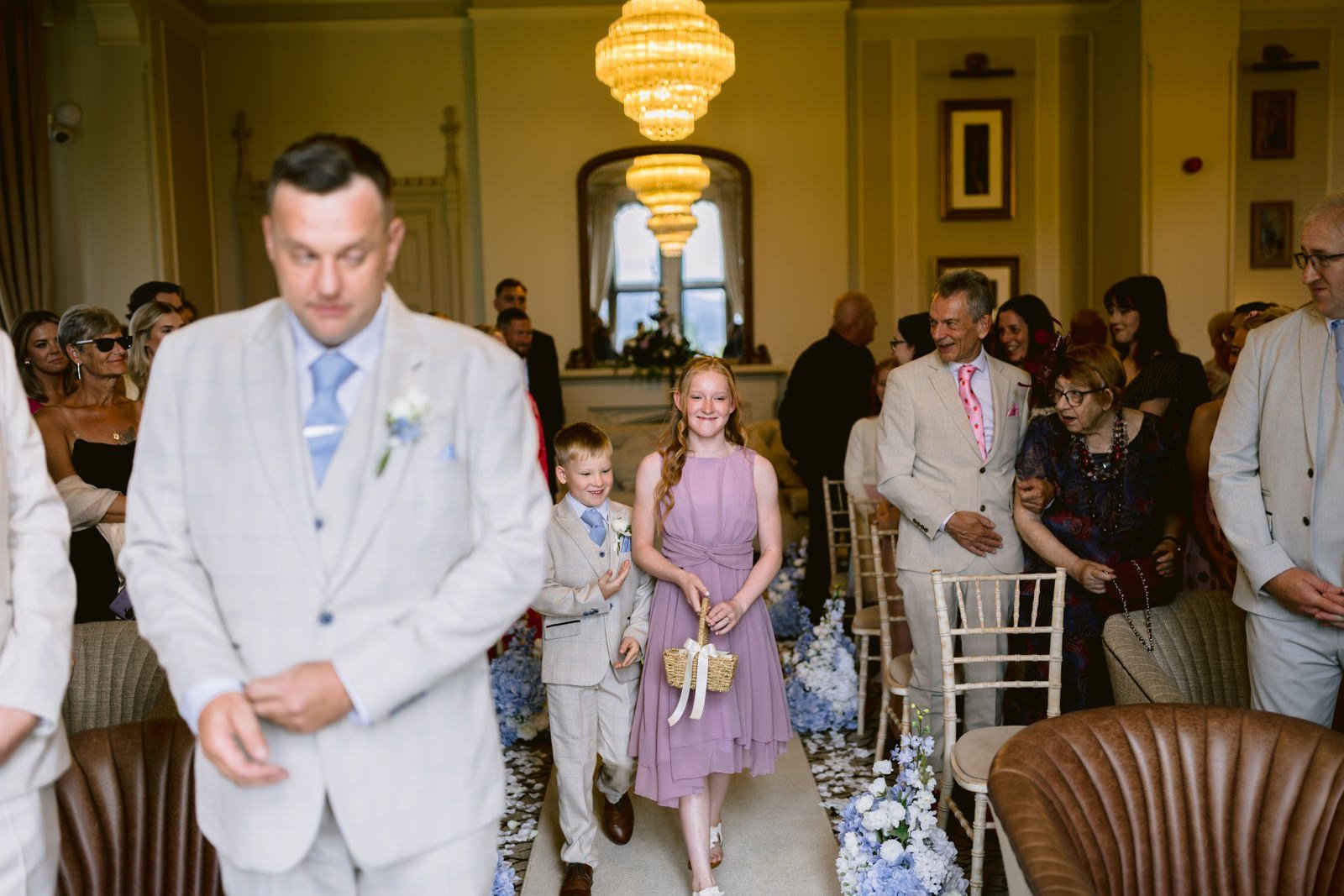 Children walk down an aisle sprinkled with flower petals, smiling, as guests seated on both sides watch during a formal August Summer Wedding at Armathwaite Hall.