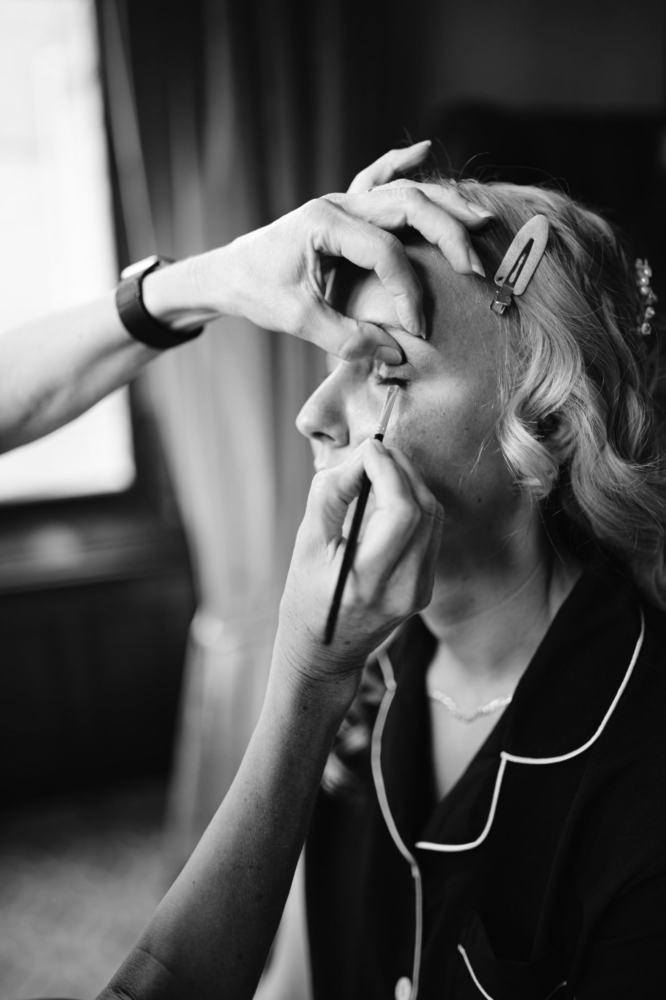 A person applies eye makeup to a woman with curled hair, dressed in contrast-piped pajamas, as she prepares for an August summer wedding at Armathwaite Hall. The image is captured in striking black and white.