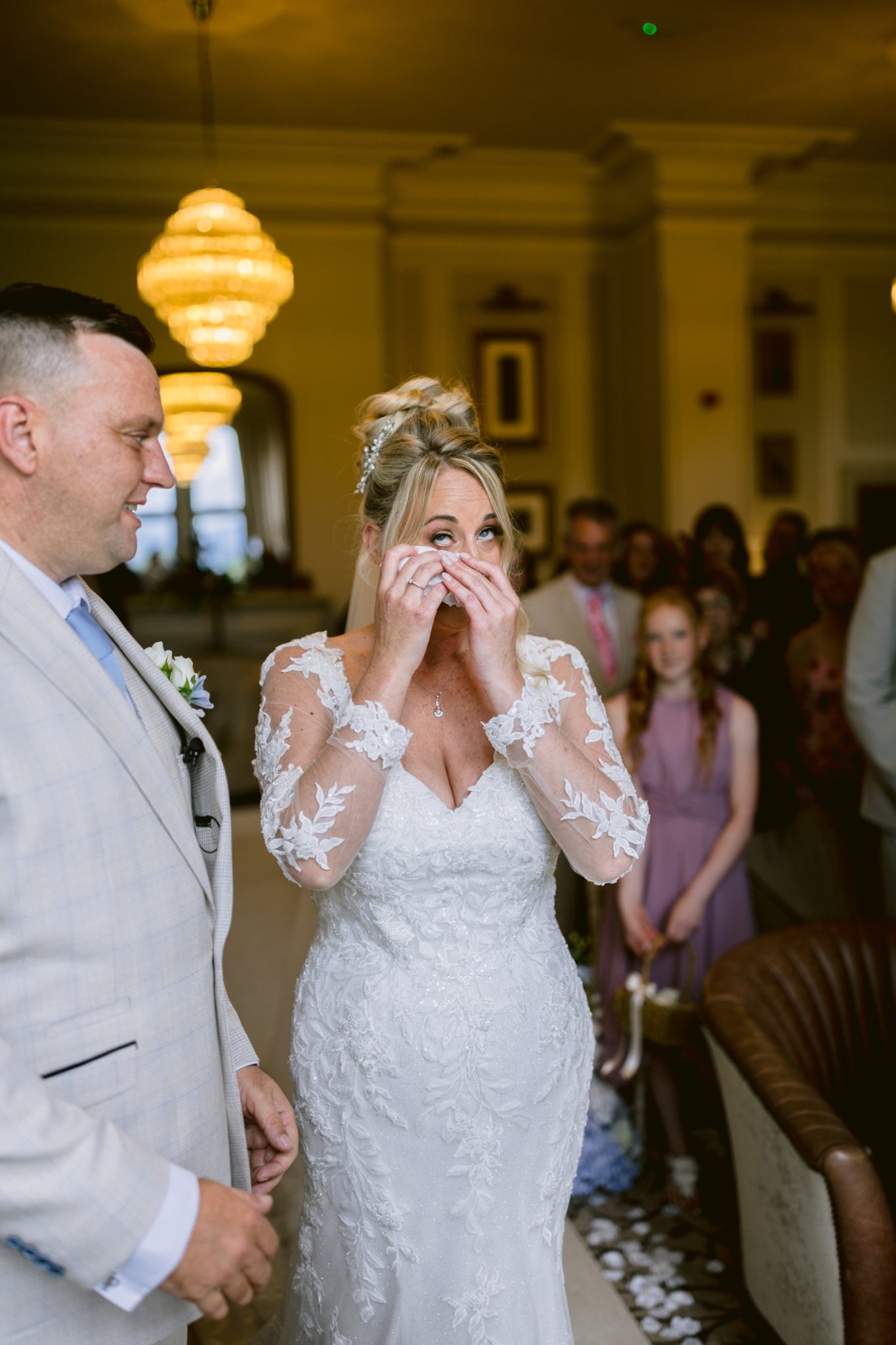A bride in a white lace dress wipes tears from her eyes, standing next to a groom in a light suit at their August Summer Wedding, surrounded by guests indoors at Armathwaite Hall.