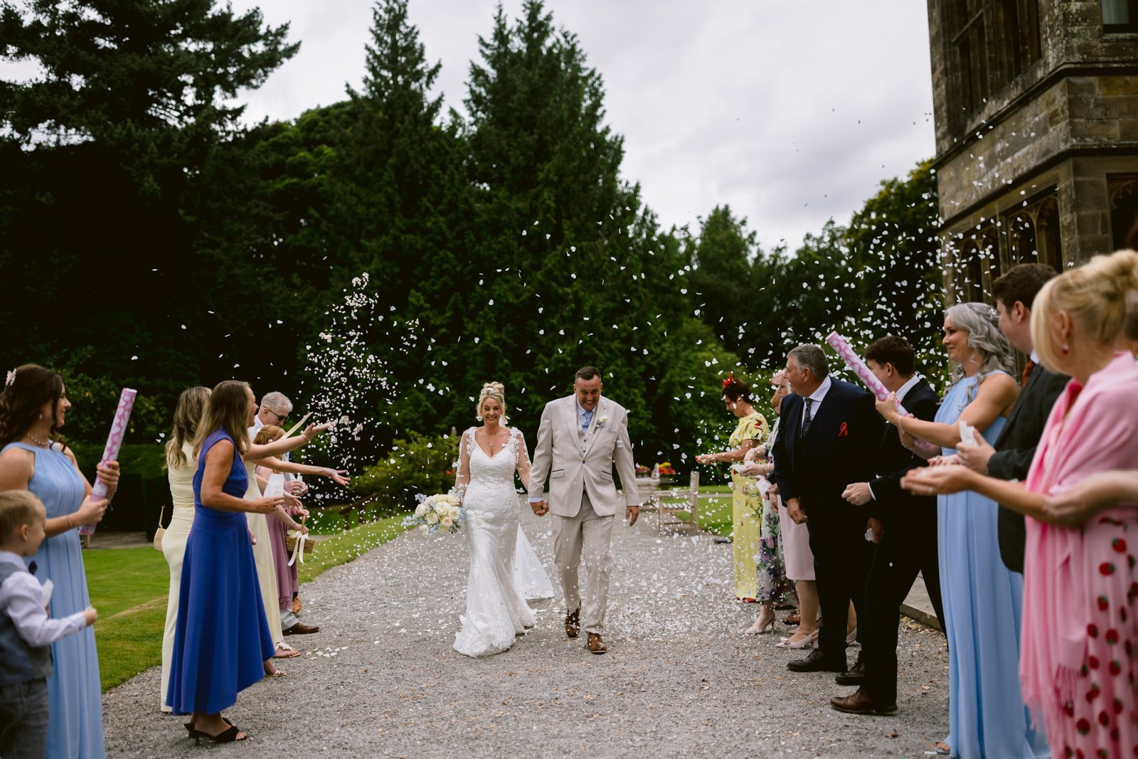 A bride and groom walk down an outdoor path at Armathwaite Hall while guests on both sides celebrate by releasing confetti, making for a joyful August Summer Wedding.