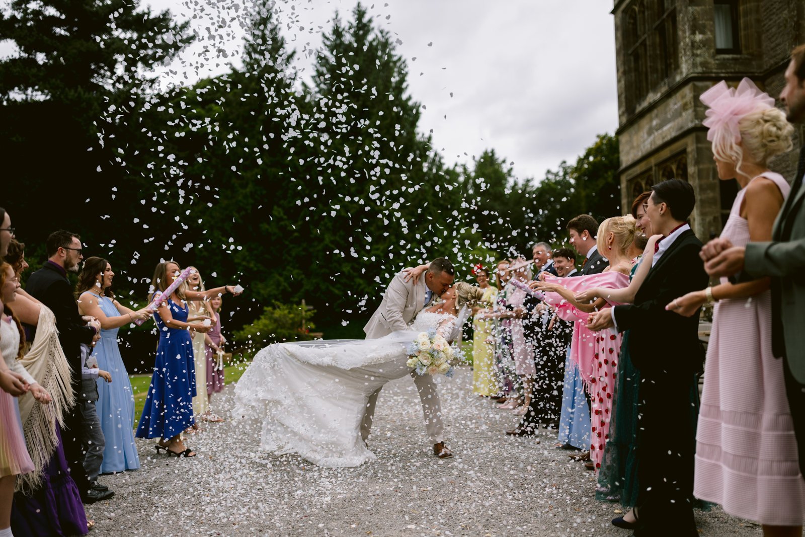 A groom dips and kisses the bride as guests in formal attire throw white confetti outdoors at Armathwaite Hall, capturing the magic of an August Summer Wedding near a historic building.