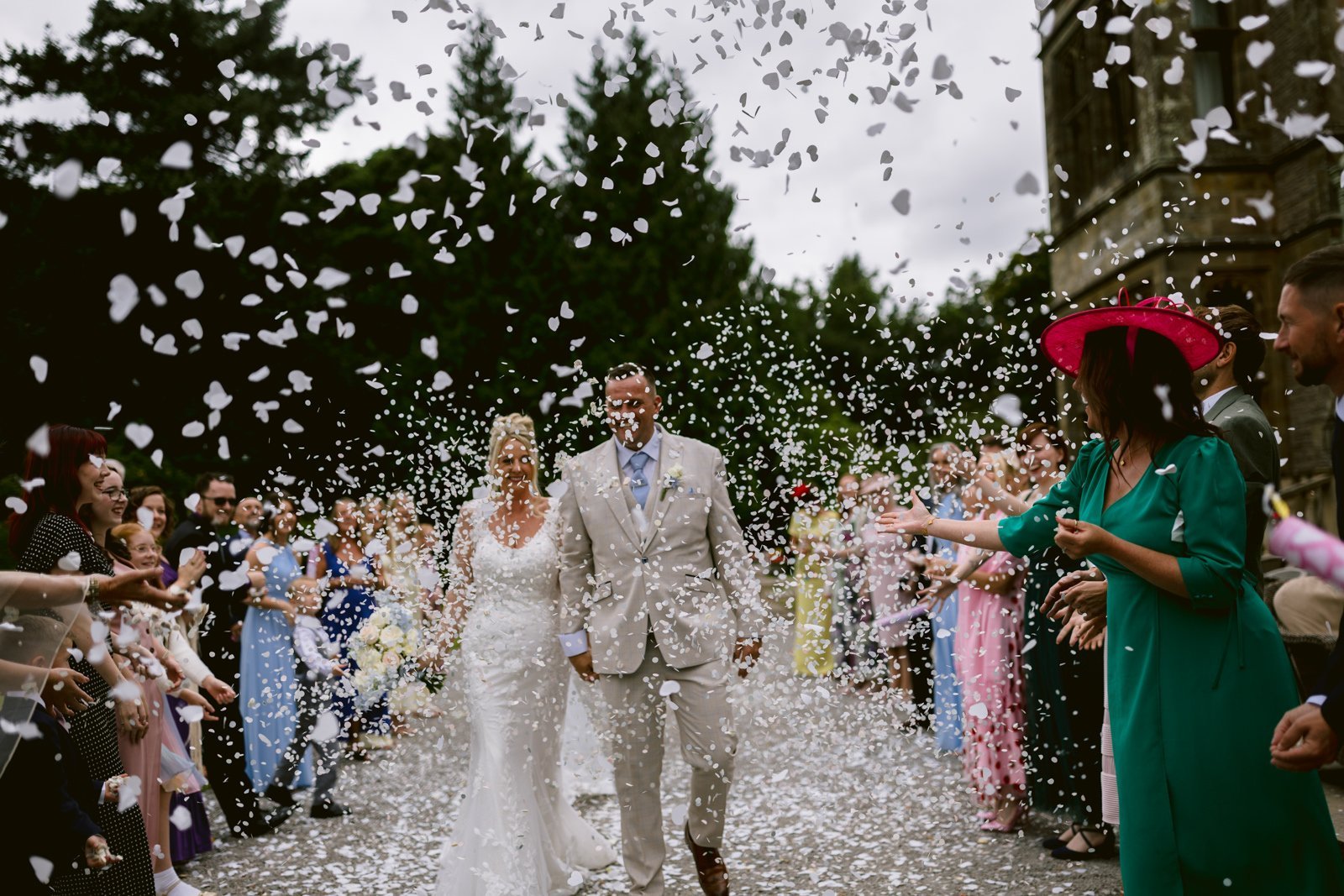 A bride and groom walk outdoors at Armathwaite Hall, surrounded by guests throwing white confetti beneath August summer wedding skies, with trees and a building in the background.