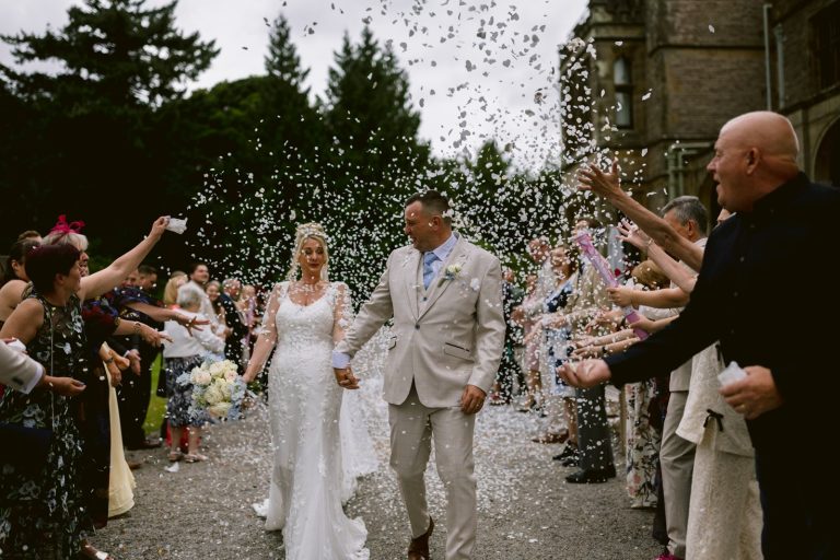 The bride and groom walk hand in hand outside Armathwaite Hall while guests throw confetti, celebrating their summer wedding ceremony.