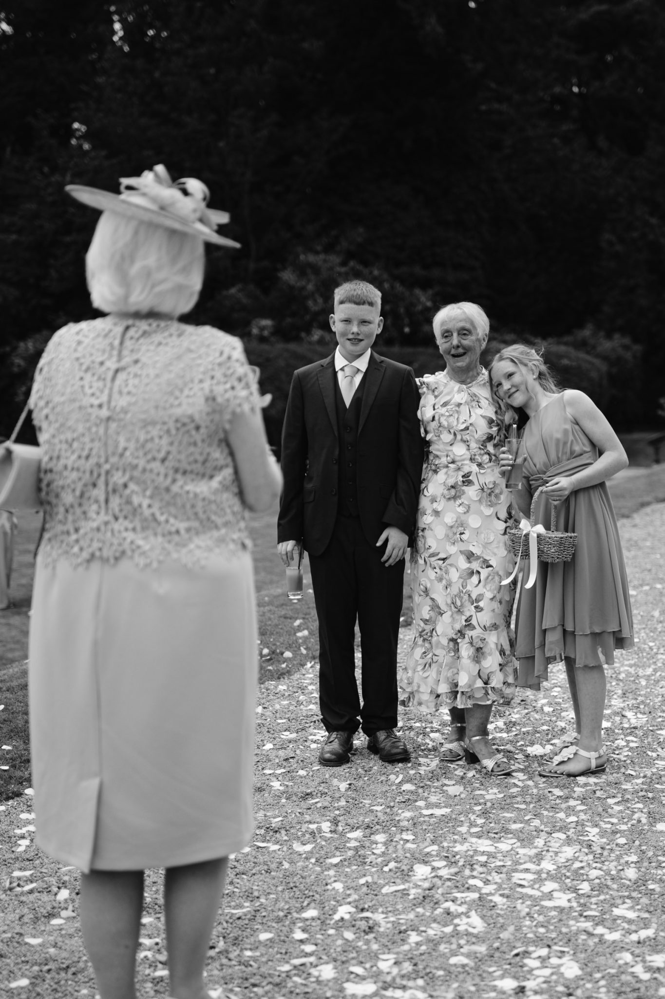 Three people pose for a photo outdoors at Armathwaite Hall while an older woman in a hat faces them; flower petals are scattered on the ground, capturing the charm of an August Summer Wedding.