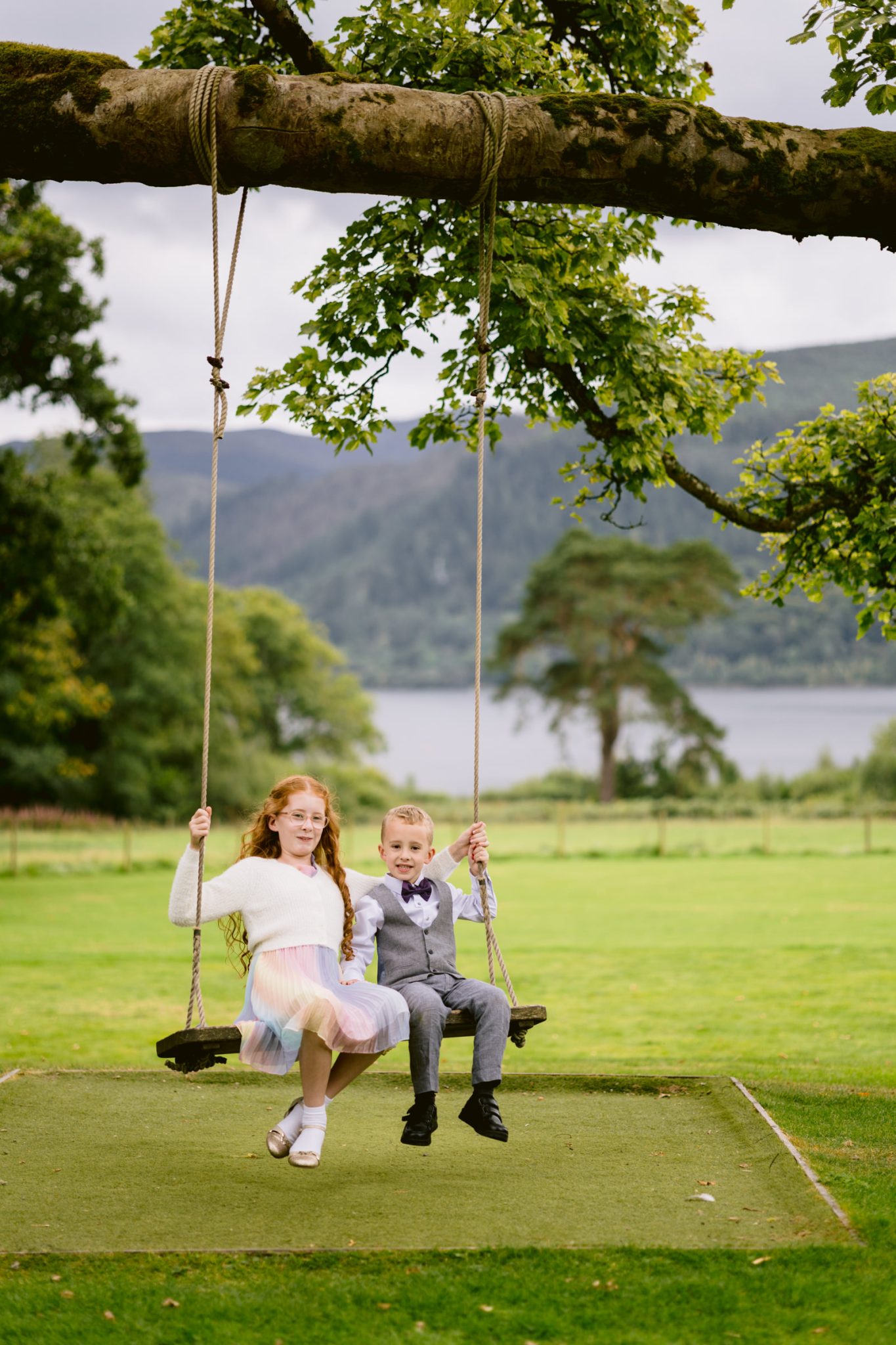 Two children, a girl in a pastel dress and a boy in a gray suit, sit together on a large wooden swing at Armathwaite Hall, enjoying an August summer wedding with trees and a lake as their picturesque backdrop.