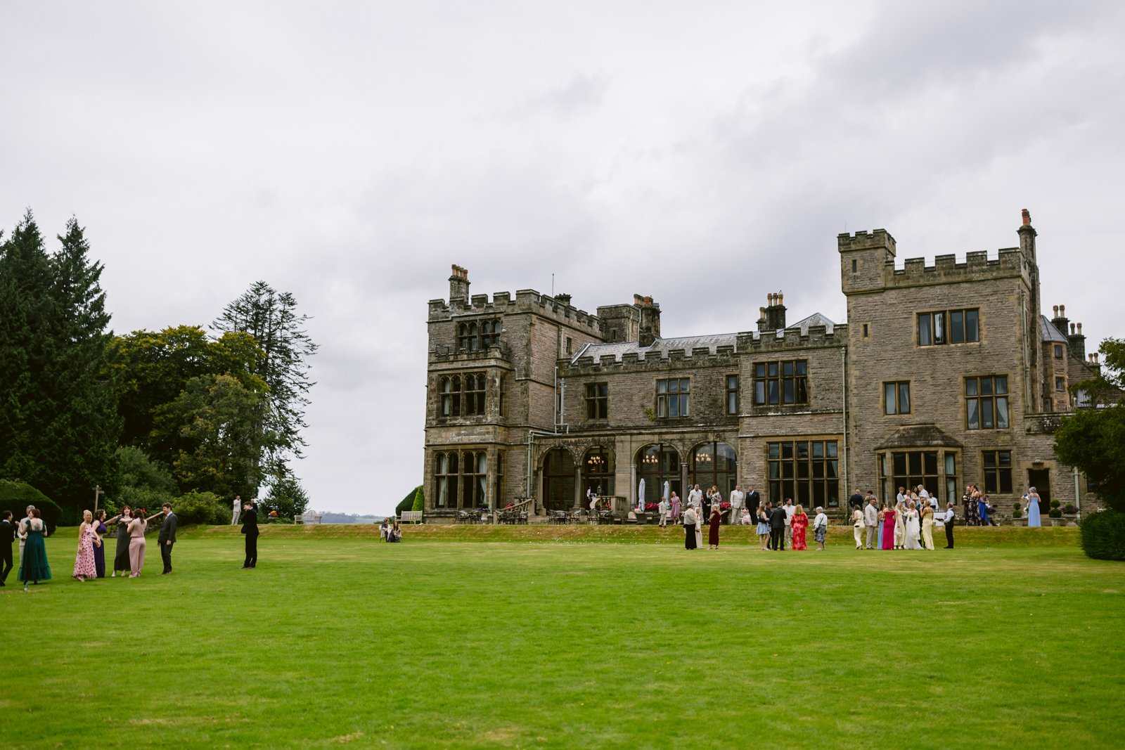 Guests in formal attire gather on the lawn outside Armathwaite Hall, a grand stone manor with turrets and tall windows, enjoying an August summer wedding beneath a cloudy sky.