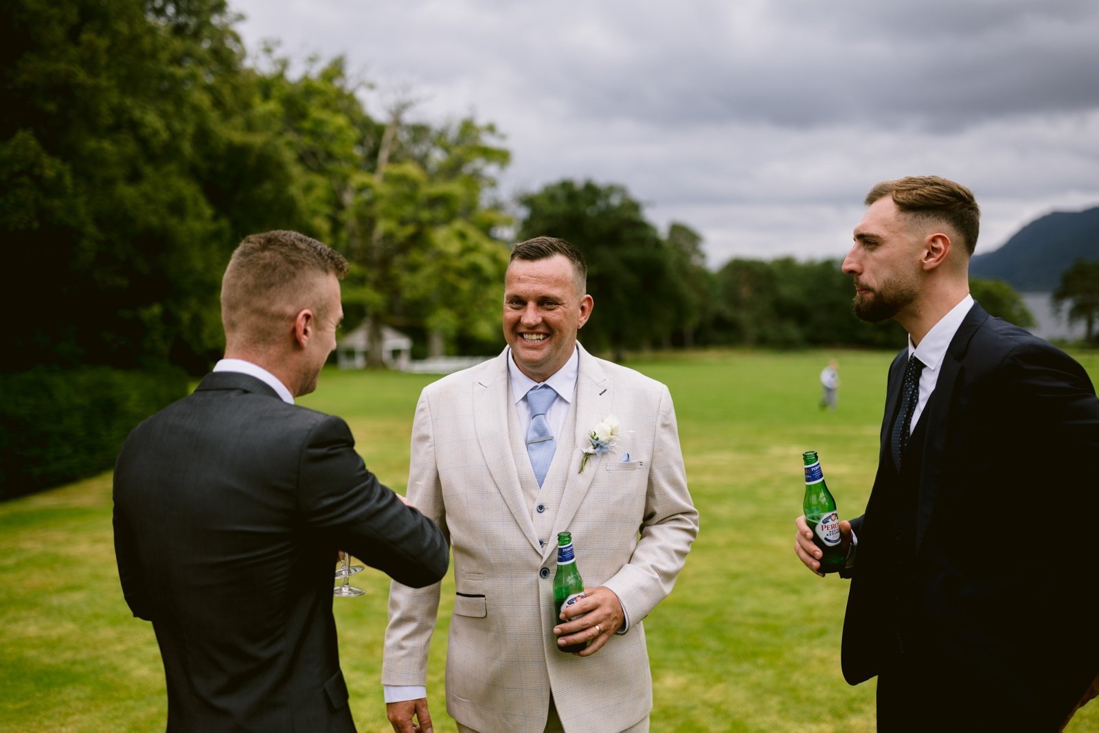 Three men in suits stand on a lawn at Armathwaite Hall, two holding bottled drinks, and converse beneath a cloudy sky during an August summer wedding.