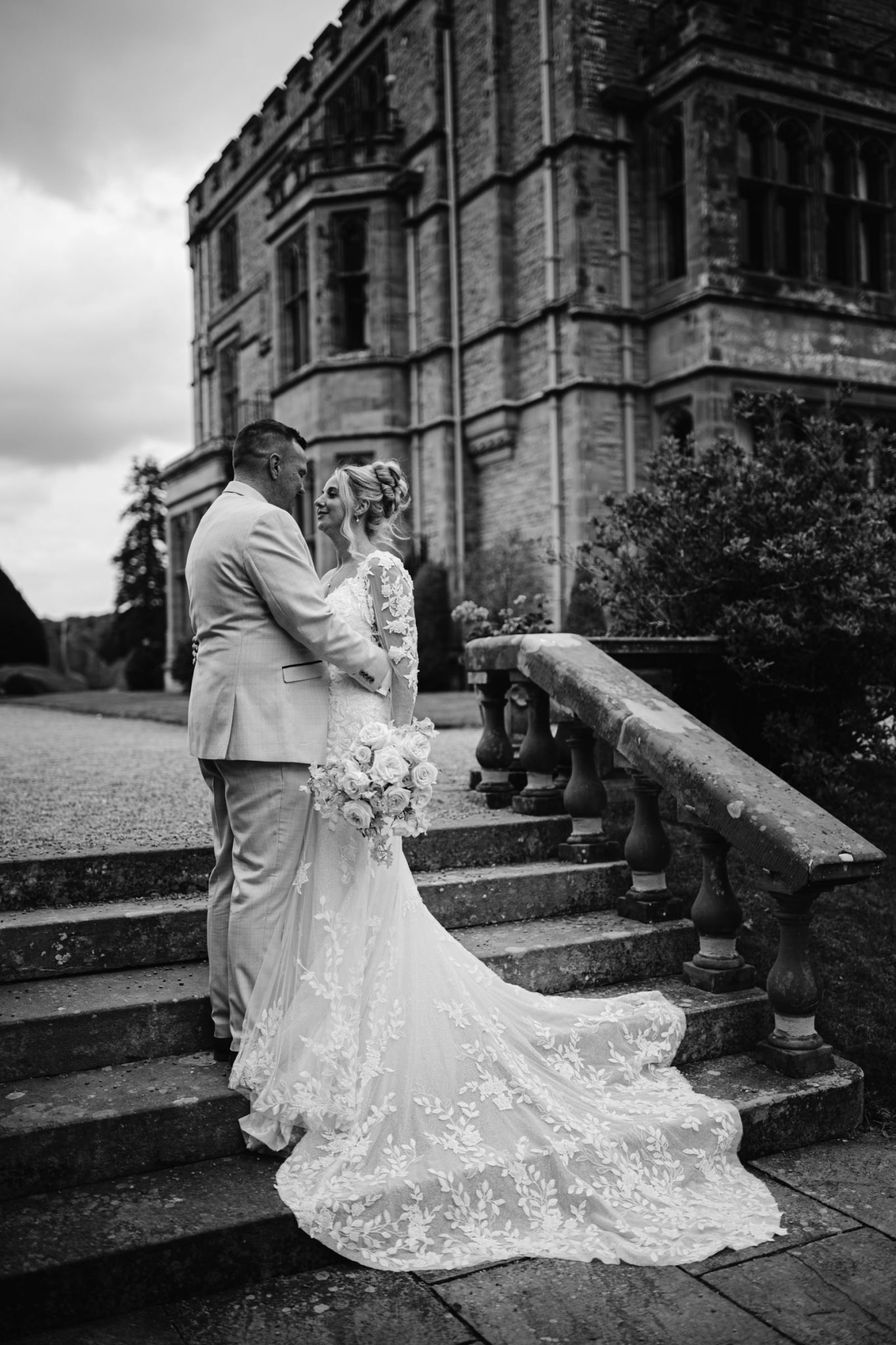 A bride and groom stand on stone steps at Armathwaite Hall, embracing and facing each other, with a bouquet in hand and the grand building behind them—capturing a perfect August Summer Wedding moment.