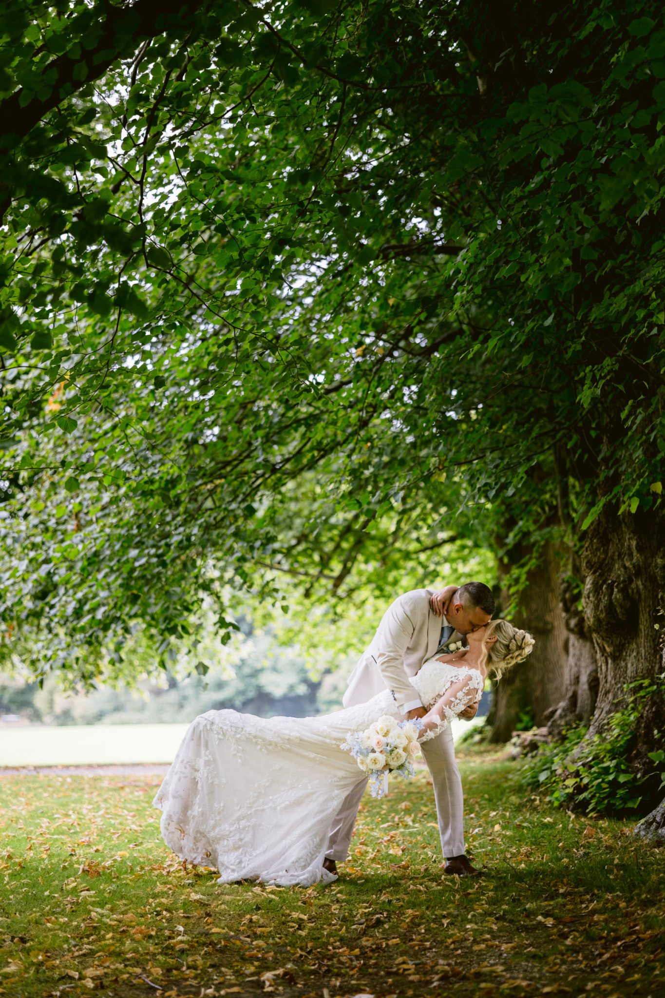 A newlywed couple dressed in formal wedding attire shares a kiss outdoors under large green trees at Armathwaite Hall, with the groom gently dipping the bride during their August summer wedding.