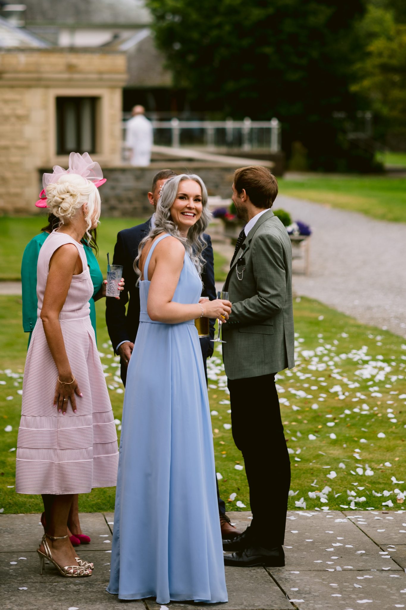 Four people dressed in formal attire stand on a garden path with scattered white petals at Armathwaite Hall; one woman in a light blue dress faces the camera and smiles, capturing the joy of an August summer wedding.