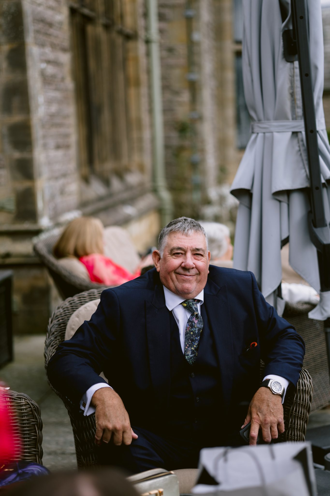 An older man in a dark suit sits in a wicker chair outdoors, smiling at the camera during an August summer wedding at Armathwaite Hall. There are people and a stone building in the background.