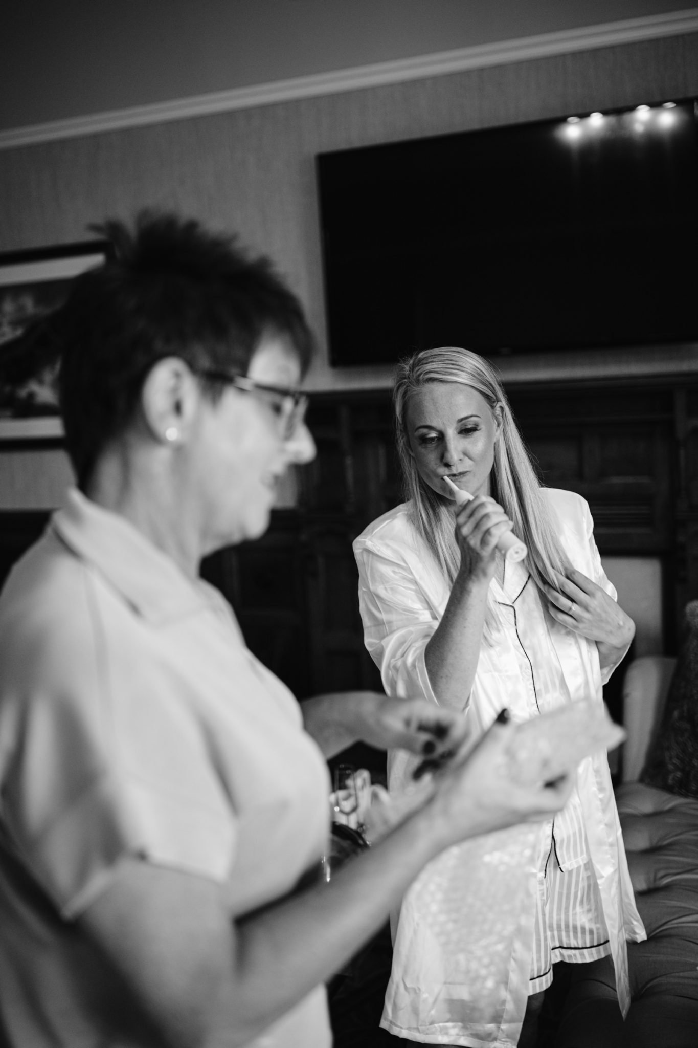 Two women indoors at Armathwaite Hall; one is brushing her teeth in pajamas, while the other is blurred in the foreground holding an object, capturing a candid moment before an August summer wedding.