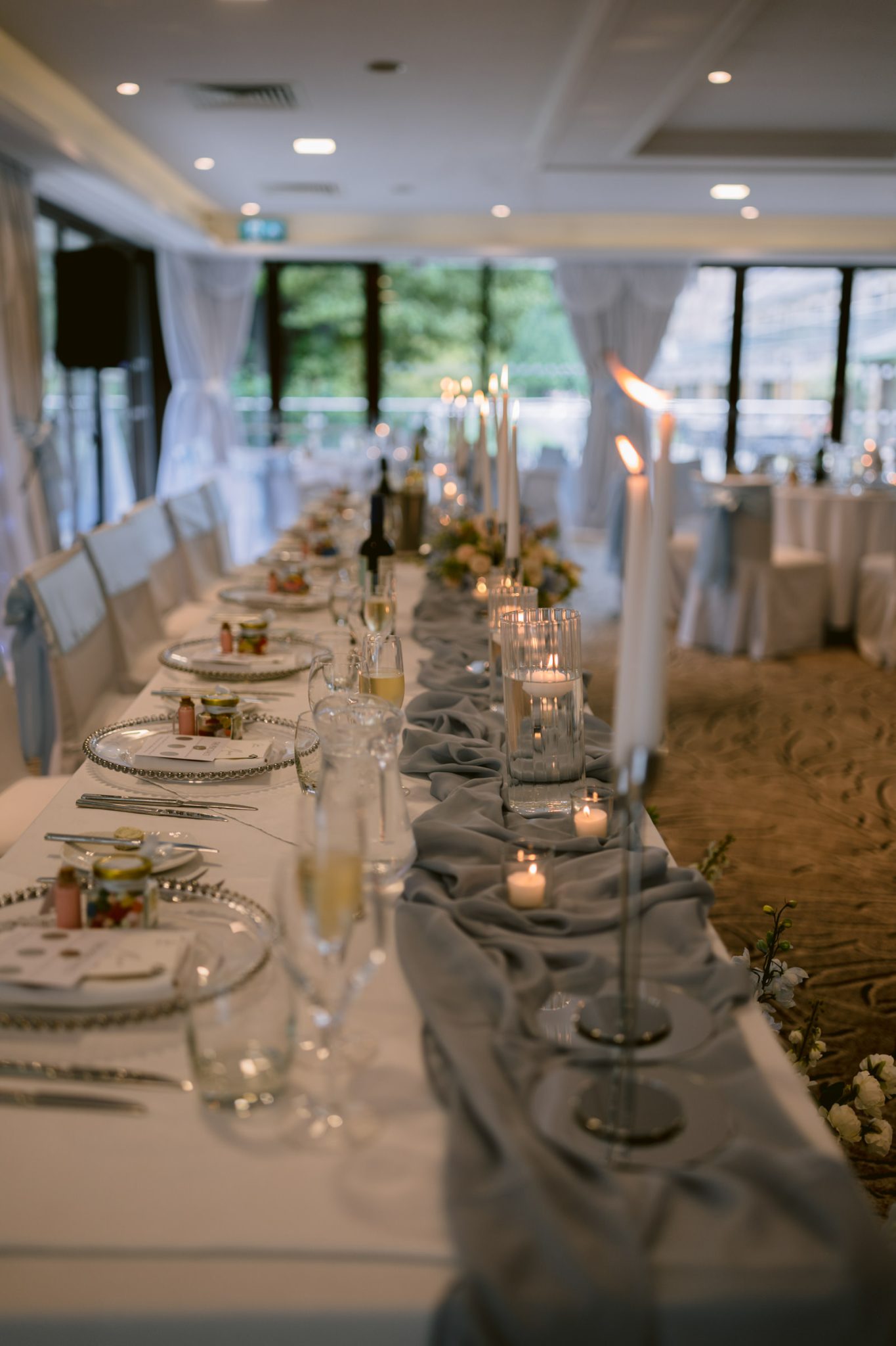 An elegantly set banquet table with white plates, glassware, candles, and a grey table runner awaits guests at Armathwaite Hall, creating a stunning scene for an August Summer Wedding in a decorated event hall with large windows.