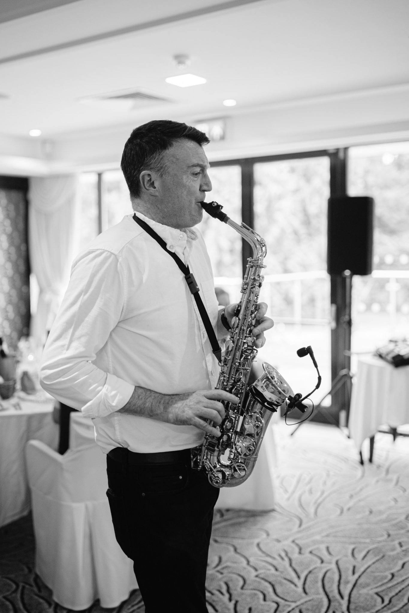 A man in a white shirt plays a saxophone indoors near tables and large windows, filling the well-lit room at Armathwaite Hall during an August Summer Wedding.
