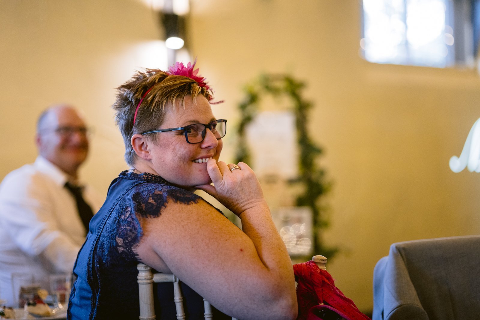 A woman in a blue lace dress and glasses sits on a chair, smiling and looking to the side at an indoor LGBT+ friendly wedding event in Shrewsbury, with a man in the background.