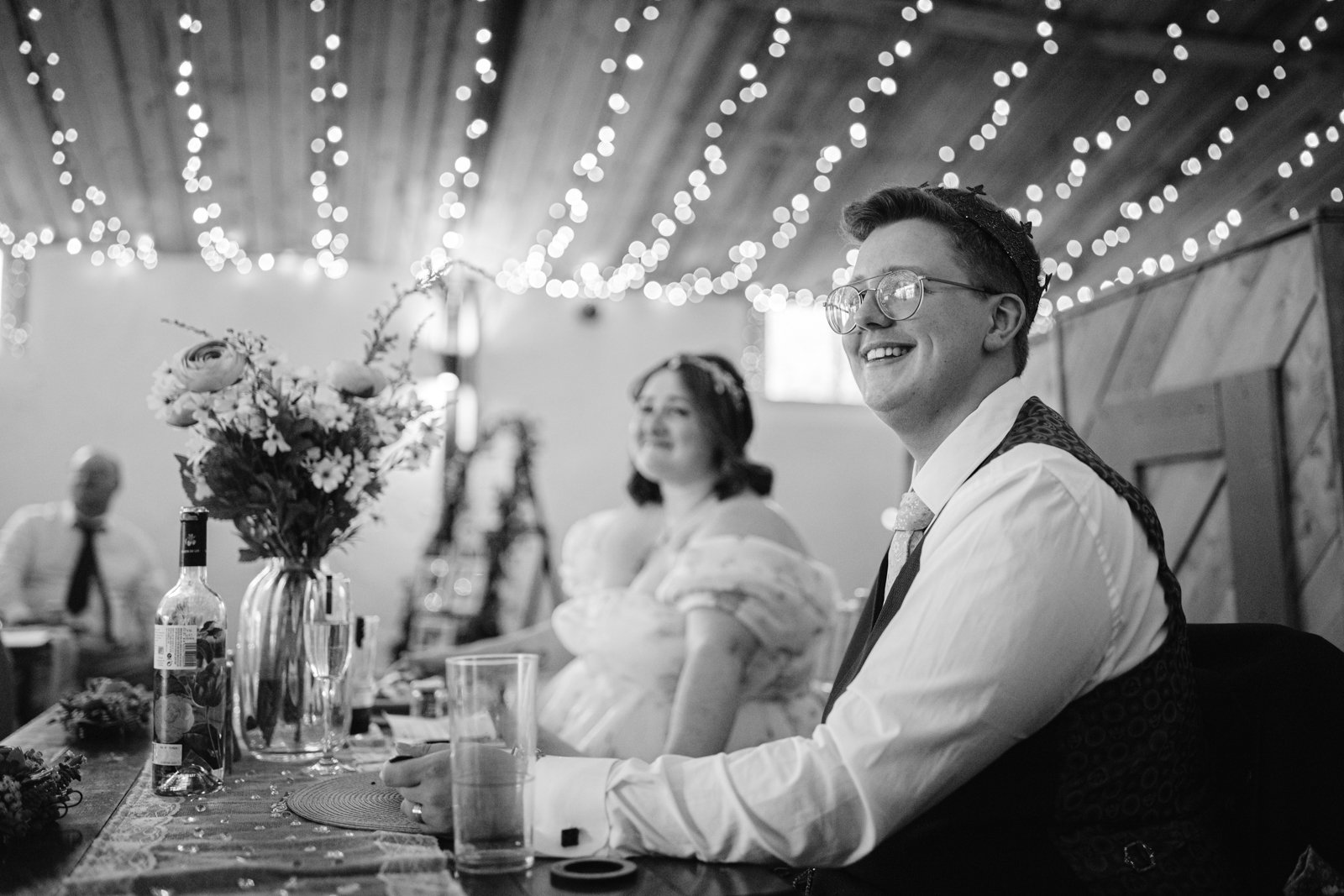 A couple dressed in wedding attire sits at a decorated table under string lights, with drinks and flowers in the foreground—perfect for a rustic farm wedding or an LGBT+ friendly celebration.
