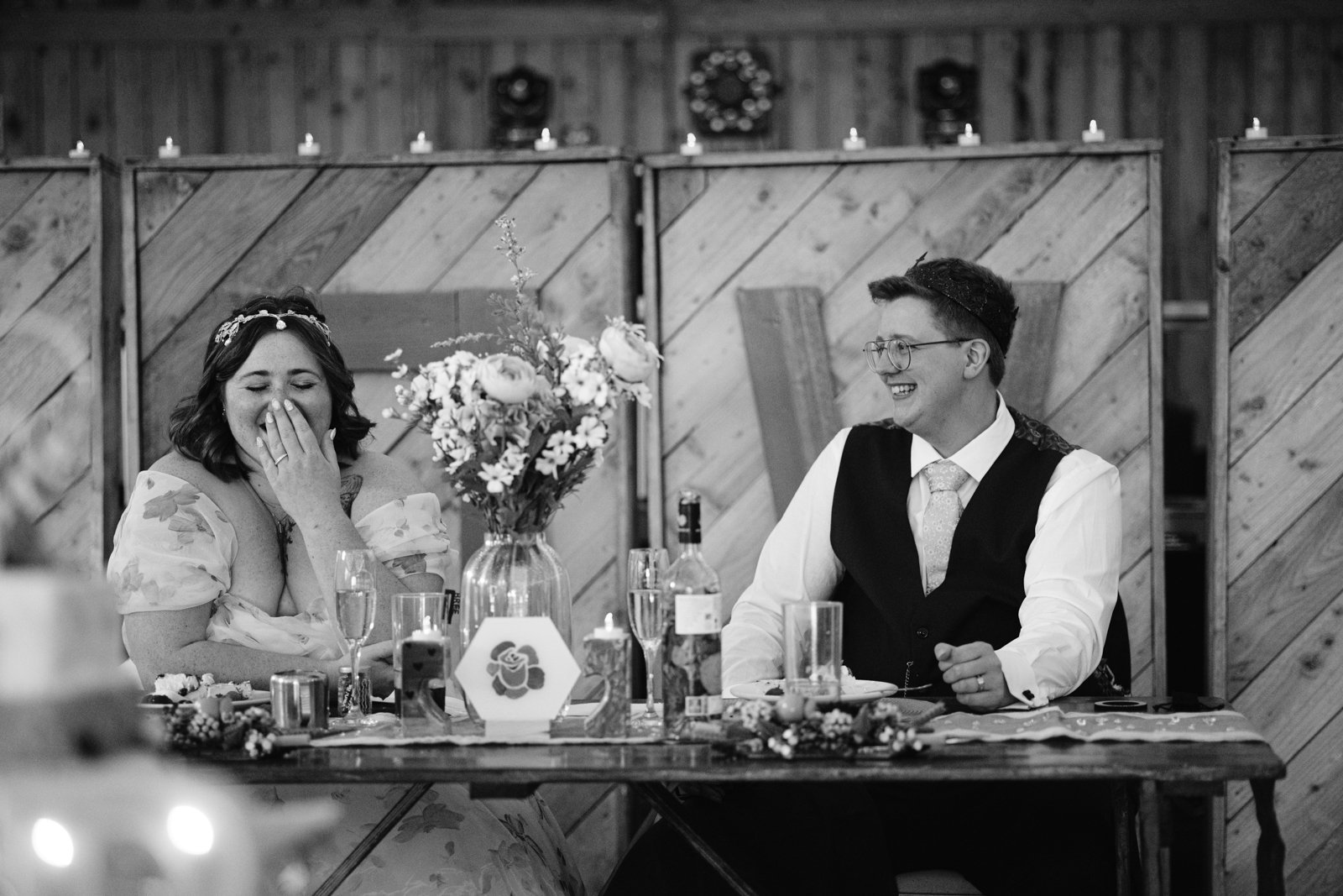 A woman and a man sit at a decorated table, both laughing. The woman has her hand to her mouth, and the man is smiling, wearing glasses and a vest. The festive scene captures the joy of an LGBT+ Friendly Wedding at rustic Stanford Farm.