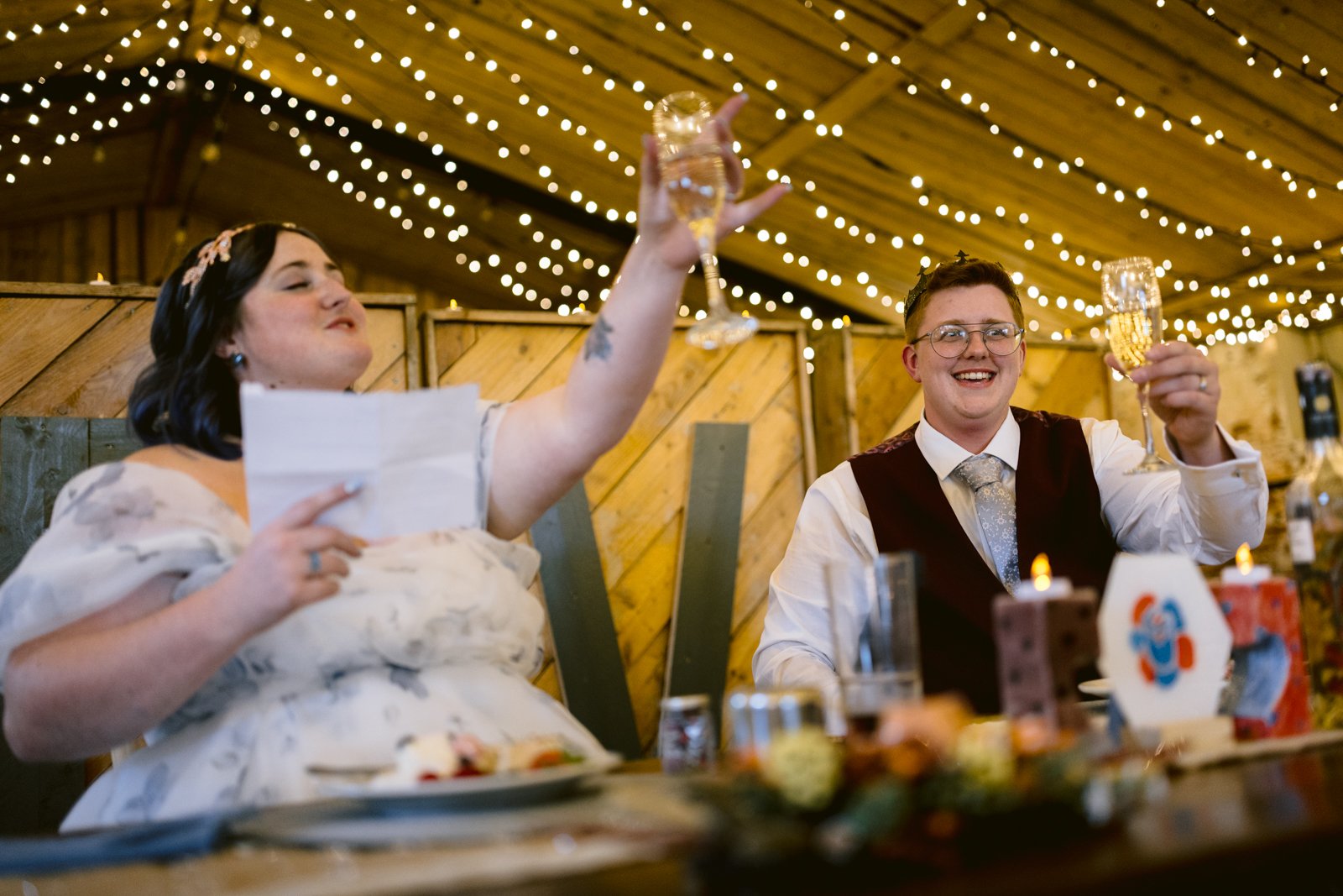 Two people in formal attire sit at a decorated table, raising champagne glasses in a toast under string lights at a rustic Stanford Farm Wedding, celebrating love in an LGBT+ friendly venue.
