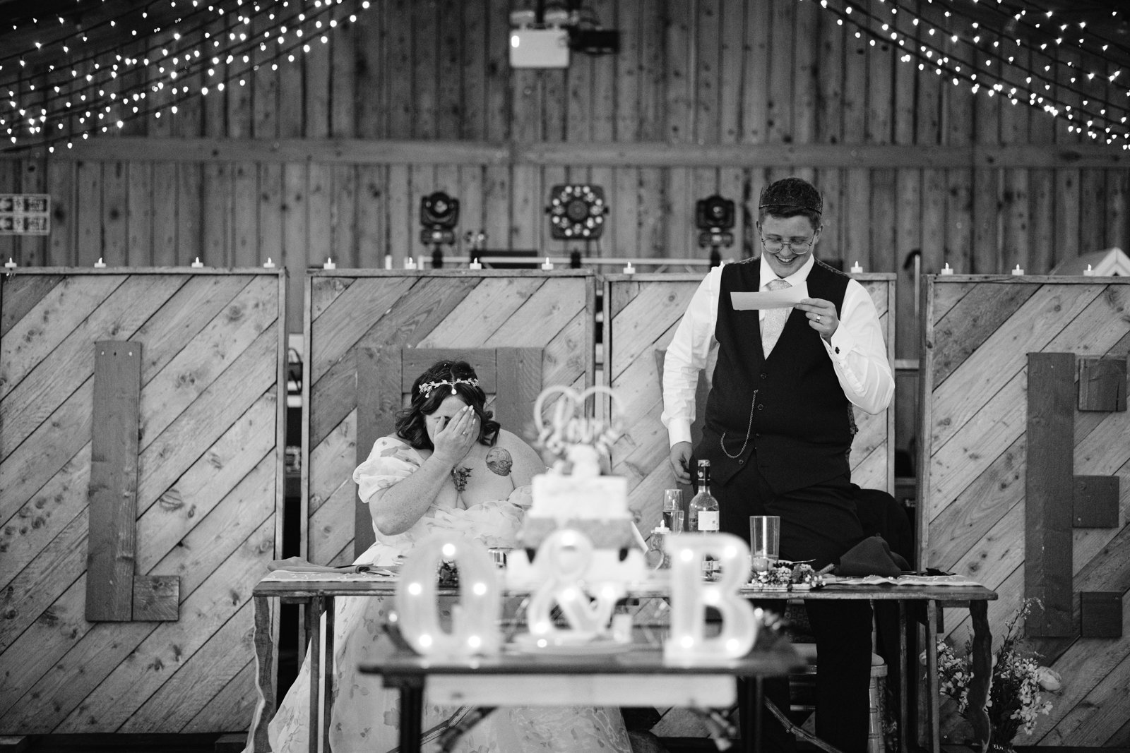 A bride wipes her eyes while a groom reads from a paper at a decorated table during an LGBT+ friendly wedding reception in a rustic venue with string lights.