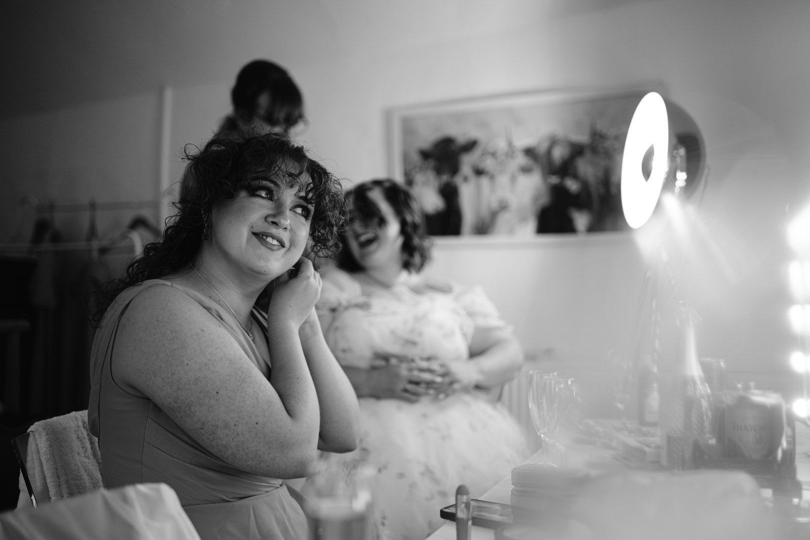 A woman in a dress adjusts her earring in front of a lighted mirror at a Stanford Farm Wedding near Shrewsbury, while another woman sits behind her, smiling, capturing the joy of an LGBT+ Friendly Wedding.