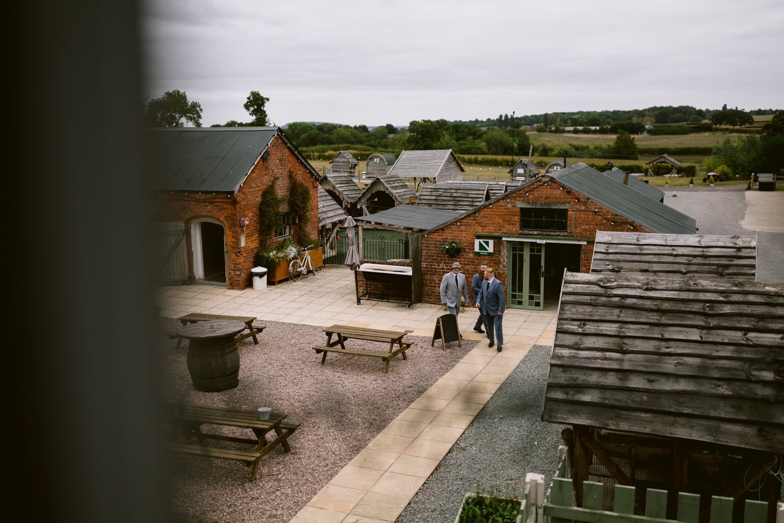Two people walk on a paved path between brick buildings in a rural courtyard, perfect for a rustic wedding or LGBT+ friendly wedding, with picnic tables and lush greenery in the background.