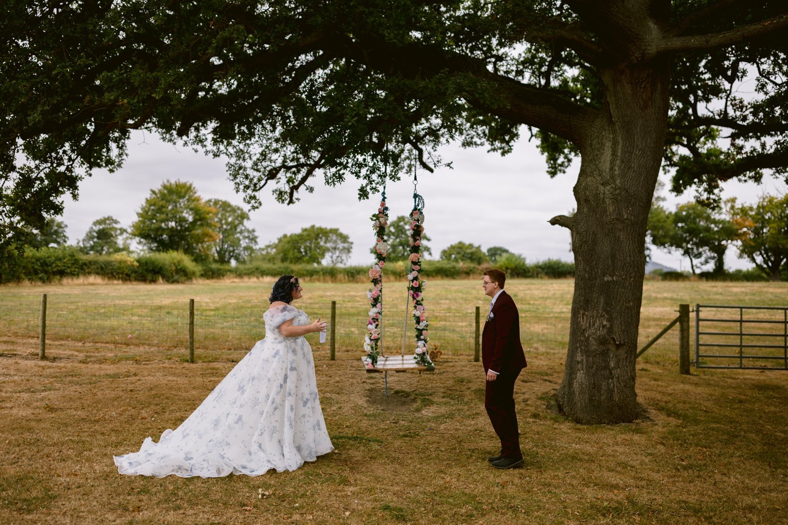 A bride in a white dress and a groom in a dark suit stand near a flower-decorated swing under a large tree in an open field at their charming Shrewsbury wedding.