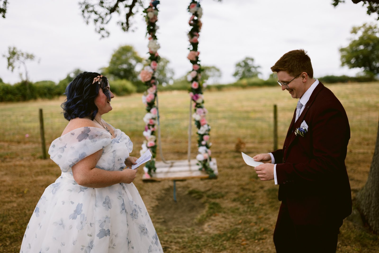 A bride and groom stand outdoors holding papers, facing each other and smiling in front of a decorated floral swing at their rustic wedding.