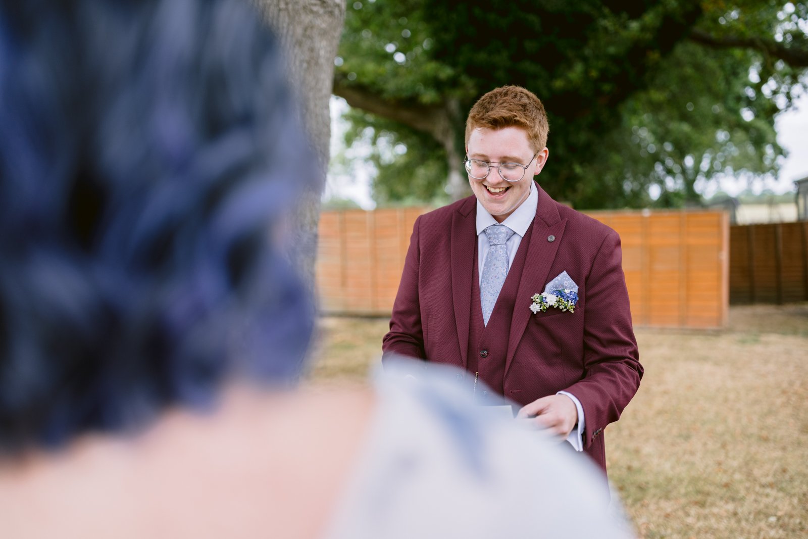 A man in a maroon suit with a boutonnière smiles outdoors at a rustic farm wedding, holding papers. A person with blue hair is visible in the foreground, out of focus.
