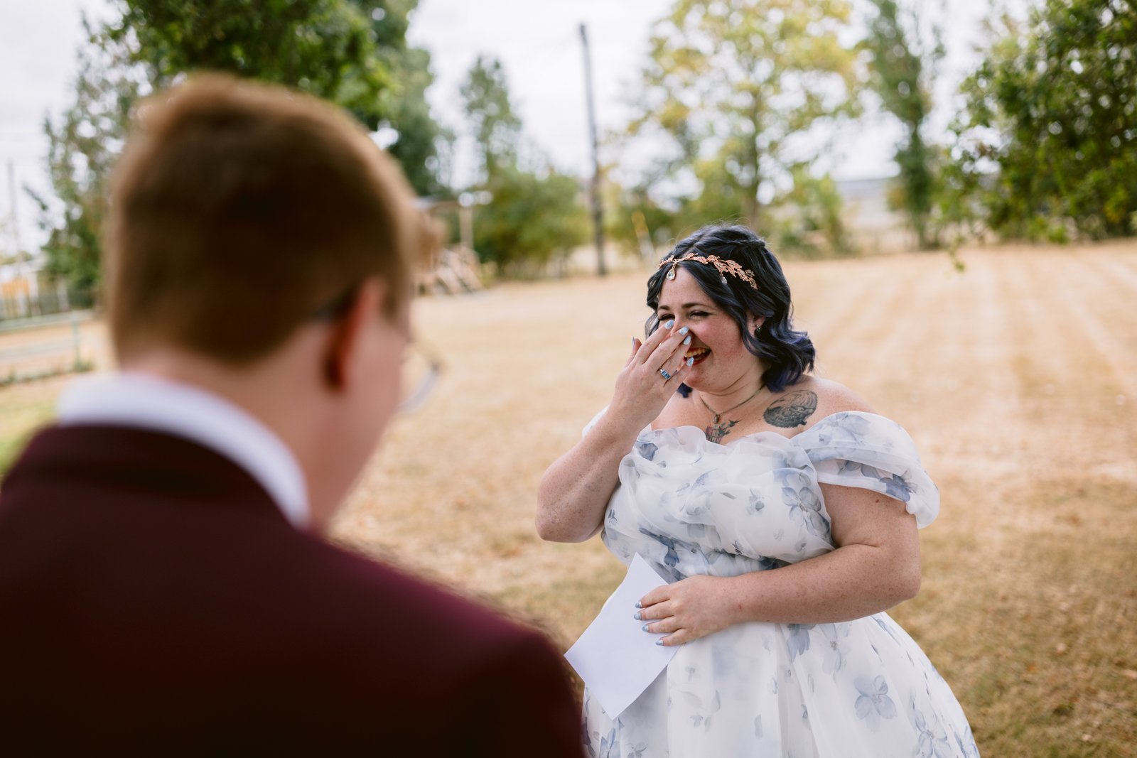 At a rustic farm wedding in Shrewsbury, a woman in a white floral dress stands outdoors, smiling and covering her mouth while facing her partner in a maroon suit. Trees and open fields set the perfect scene for this LGBT+ friendly celebration.
