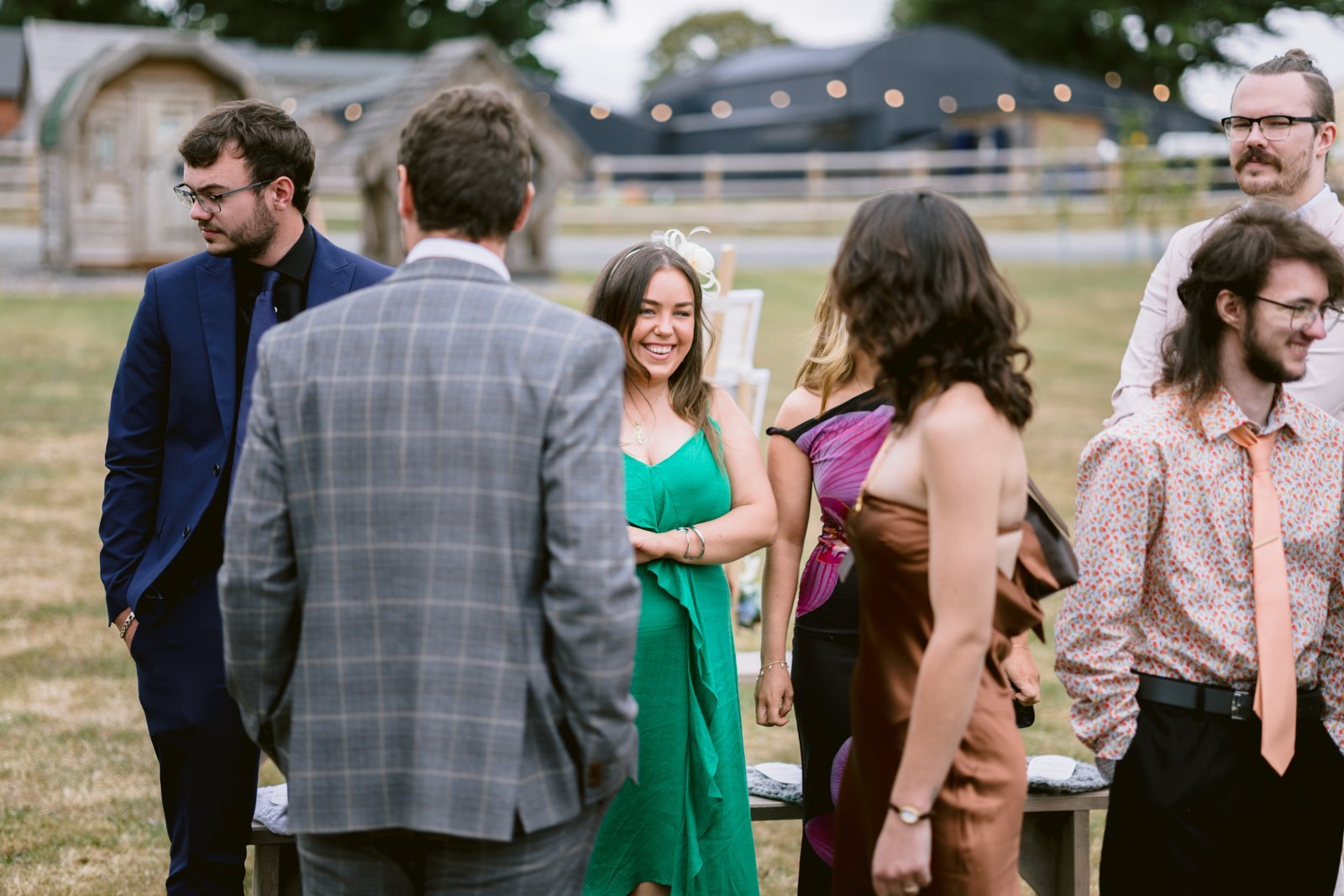 A group of people dressed in semi-formal attire stand together outdoors, talking and smiling at a rustic farm wedding, with buildings and string lights in the background.