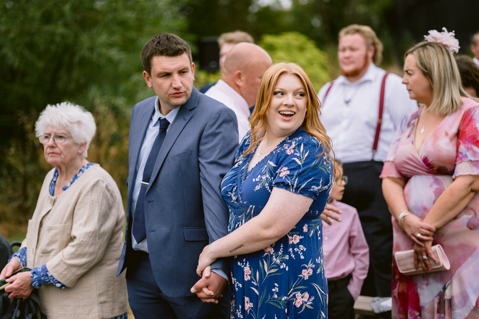 A group of people, dressed in semi-formal attire, stand outdoors at a rustic farm wedding. A woman in a blue floral dress and a man in a blue suit hold hands in the foreground, celebrating love near Shrewsbury.