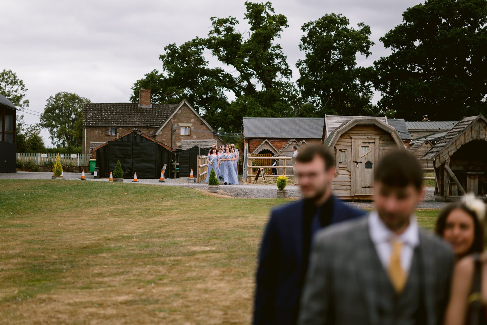 A group of bridesmaids in light purple dresses walk along a gravel path at a rustic farm wedding, while three people in suits and formal wear stand blurred in the foreground. Rustic buildings and trees complete this Shrewsbury scene.