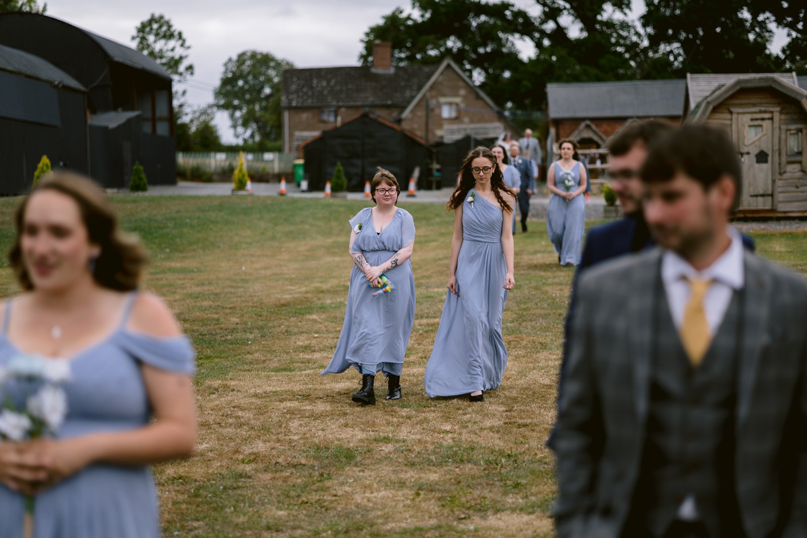 A group of people in formal attire walk outdoors on a grassy area at a rustic Stanford Farm wedding near Shrewsbury; two women in light blue dresses are in focus, with others blurred in the foreground and background.