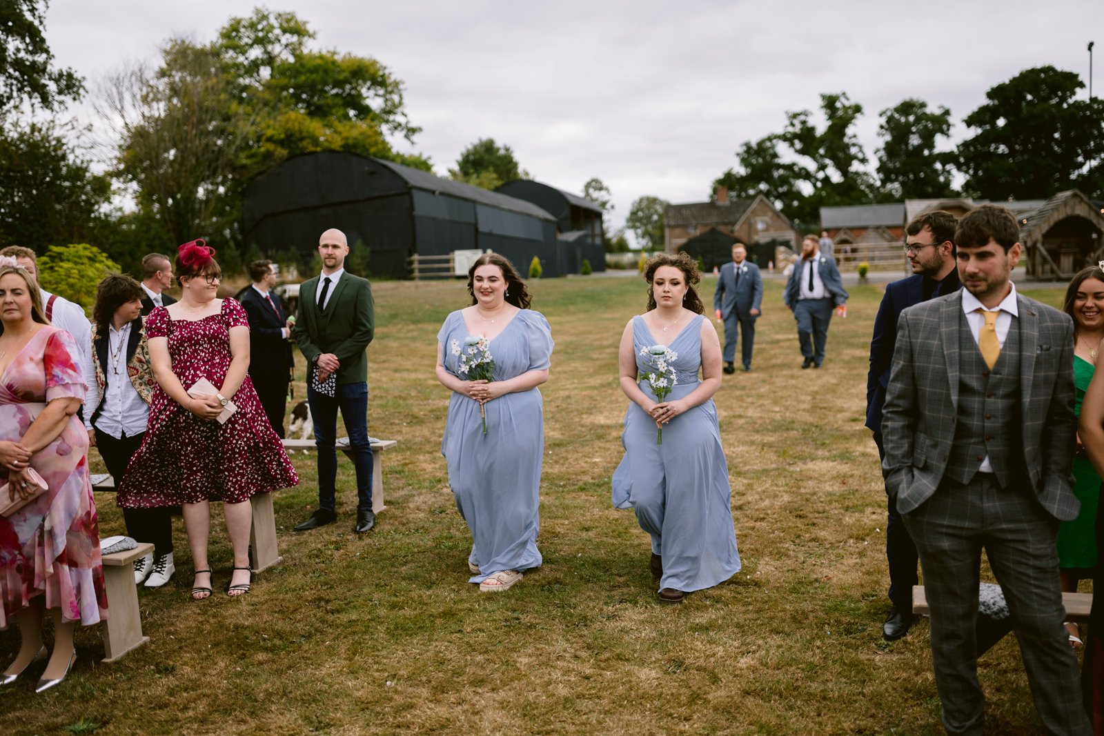 Two bridesmaids in light blue dresses walk down an outdoor aisle at a rustic Stanford Farm wedding in Shrewsbury, as guests stand on either side, with barns and trees providing a charming countryside backdrop.