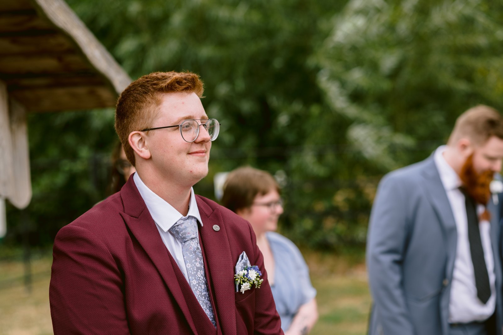 A person in a maroon suit with glasses and a floral boutonniere stands outdoors at a rustic Stanford Farm wedding near Shrewsbury, with two others in the background, one in a blue suit. The celebration is proudly LGBT+ friendly.