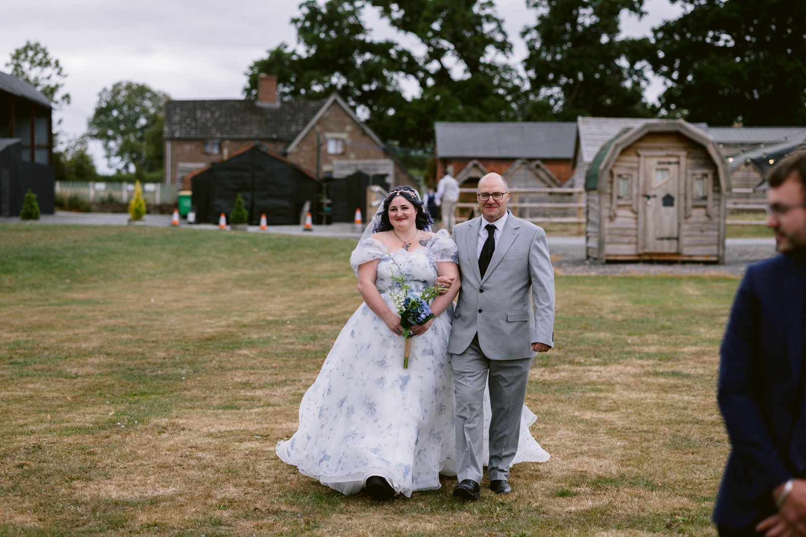 A bride in a white floral dress walks on grass holding a bouquet, accompanied by a man in a grey suit at a rustic farm wedding; charming buildings are visible in the background.