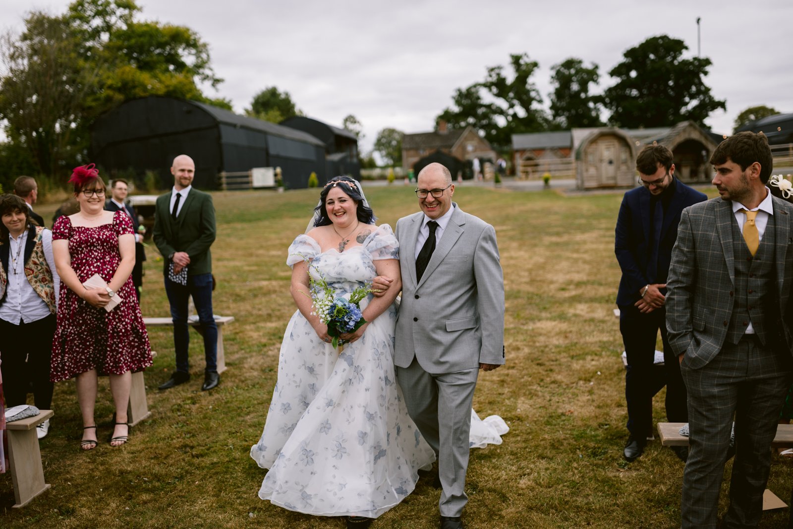 A bride in a white floral dress walks down the aisle with a man in a gray suit, surrounded by standing guests at an LGBT-friendly rustic wedding ceremony at Stanford Farm.