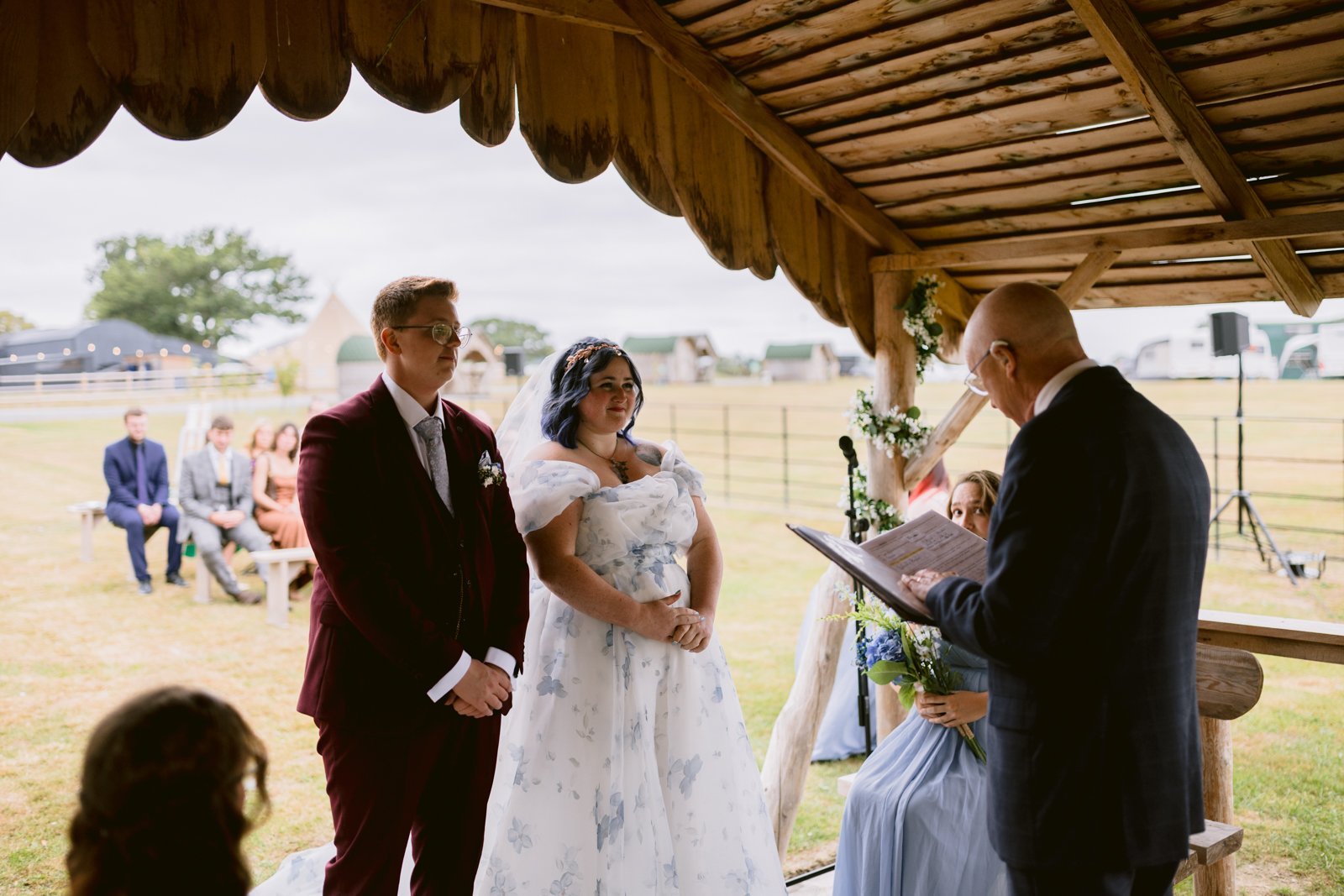 A couple stands facing an officiant under a wooden gazebo during a rustic farm wedding ceremony in Shrewsbury, with guests seated on benches in the background.